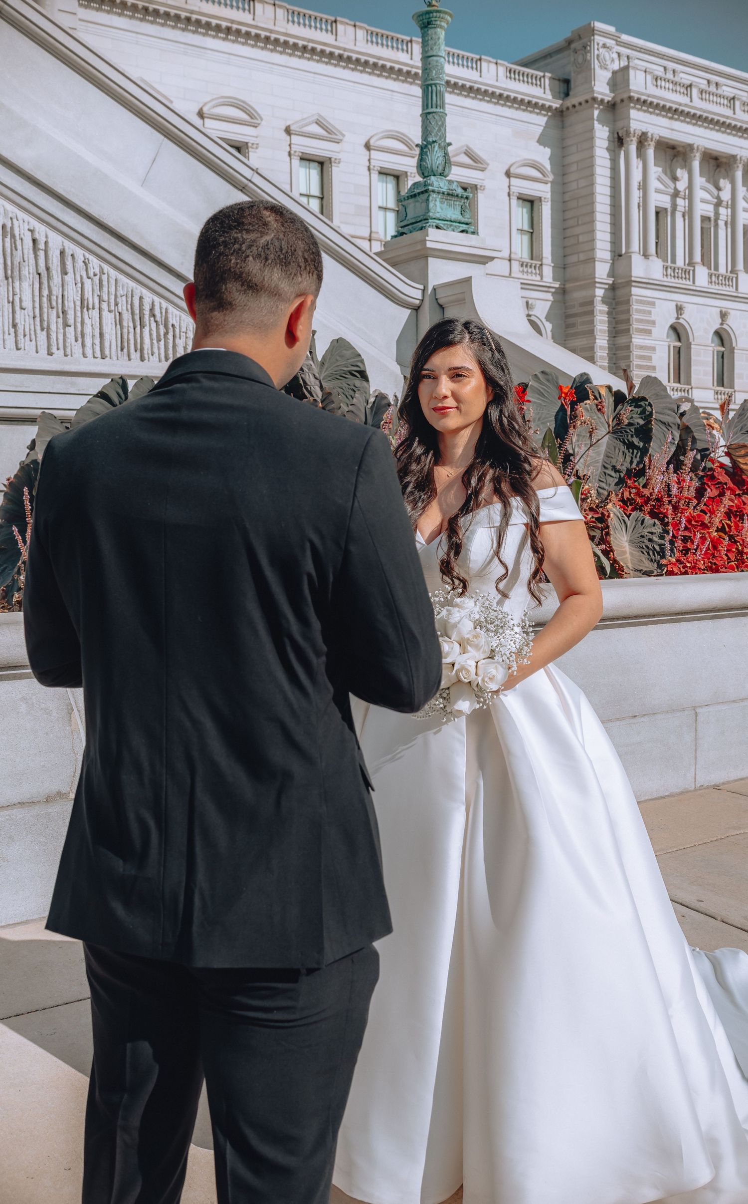 A couple dressed in wedding attire stands on the steps of a historic government building.