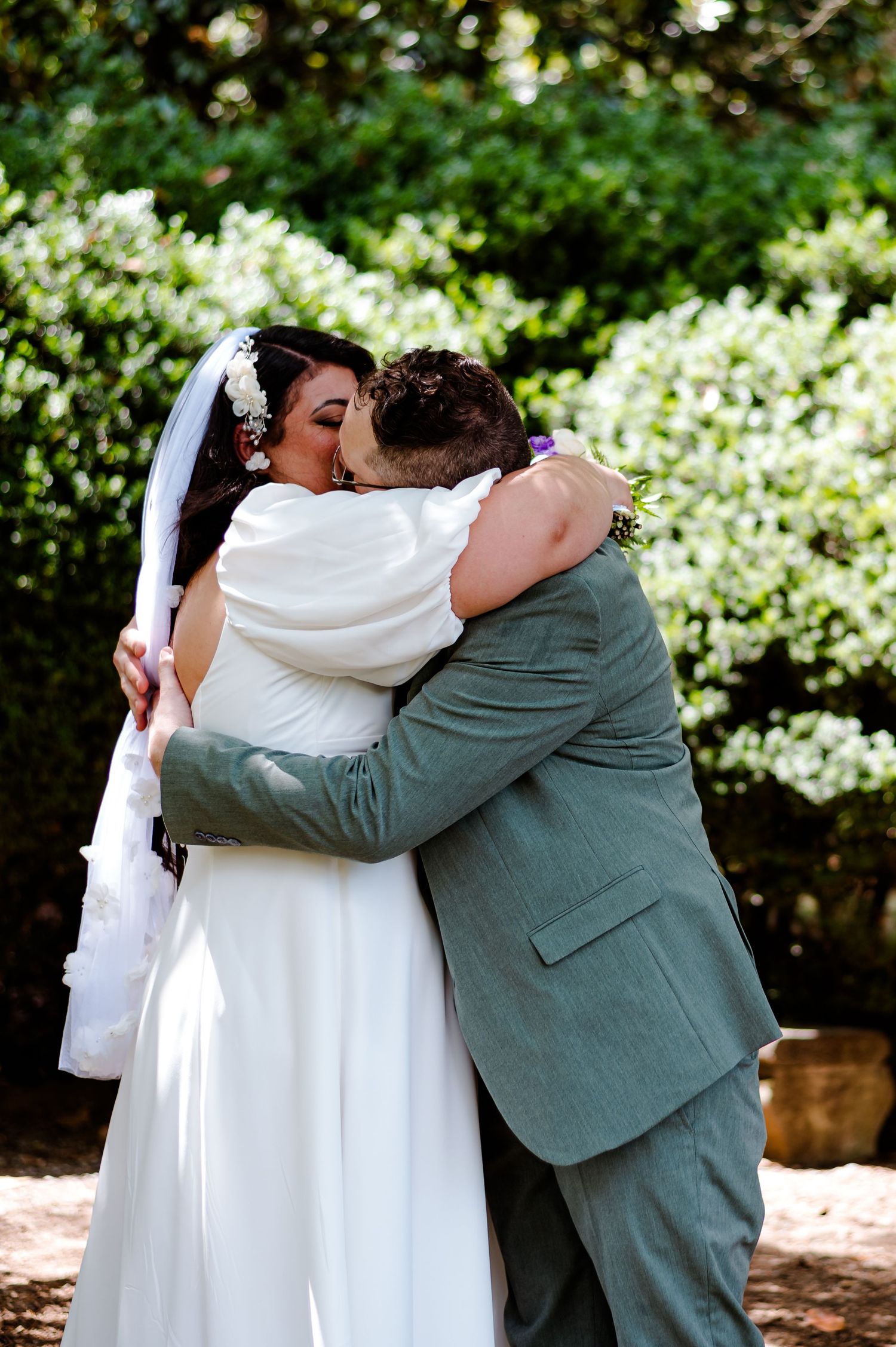 Two people embrace in a tender kiss at an outdoor wedding, with the bride wearing white and groom in a sage green suit.