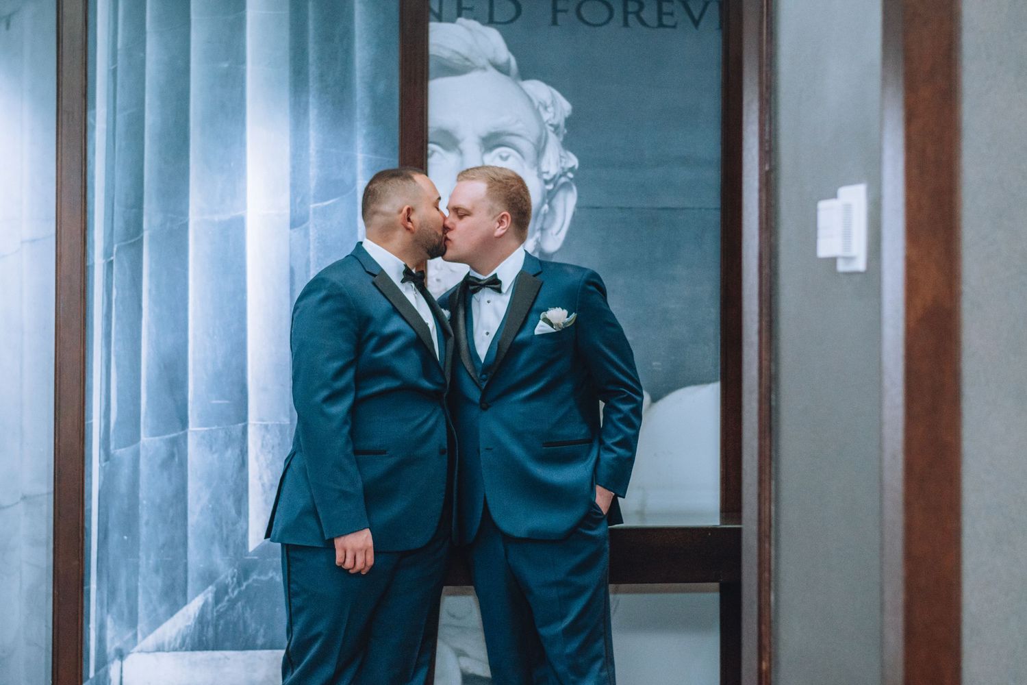 A romantic wedding moment of two people in matching navy suits sharing a kiss against a light blue building facade.