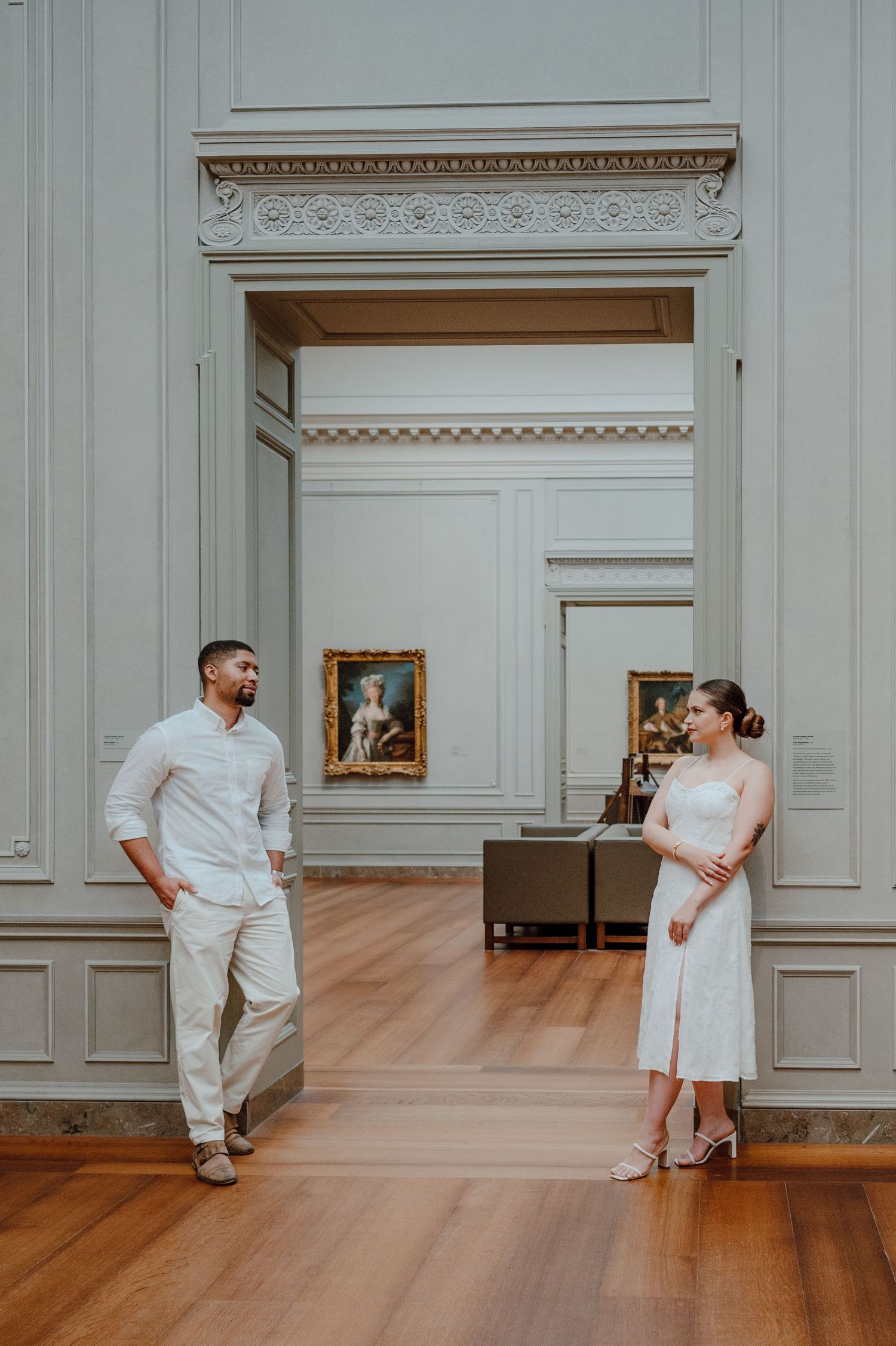 A couple in white outfits stands in an elegant museum gallery with wood floors and ornate molding on the walls.