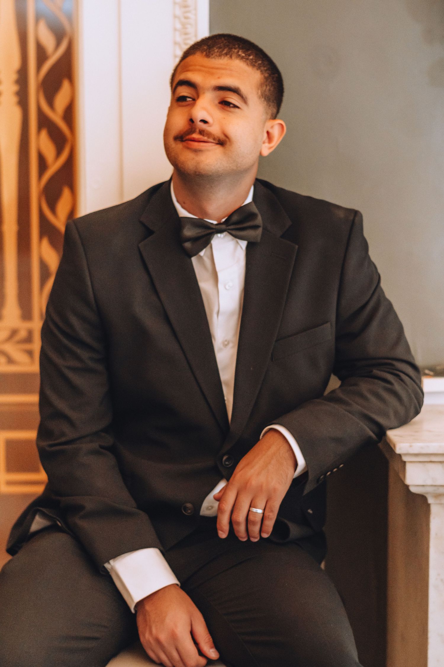A groom in black tuxedo and bowtie smiles while seated in an elegant architectural setting.