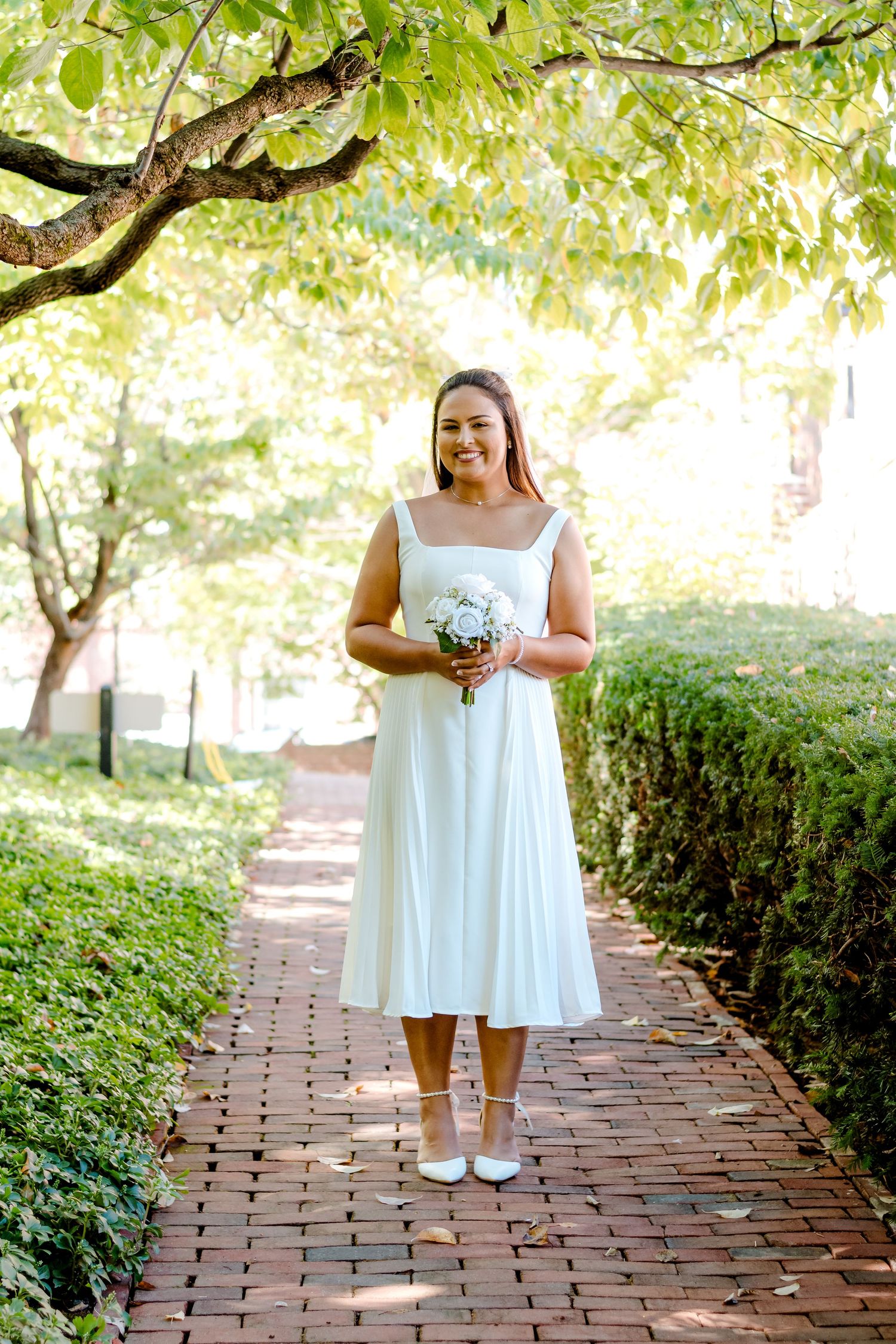 A bride in a tea-length dress stands on a brick path under green trees holding a white bouquet.