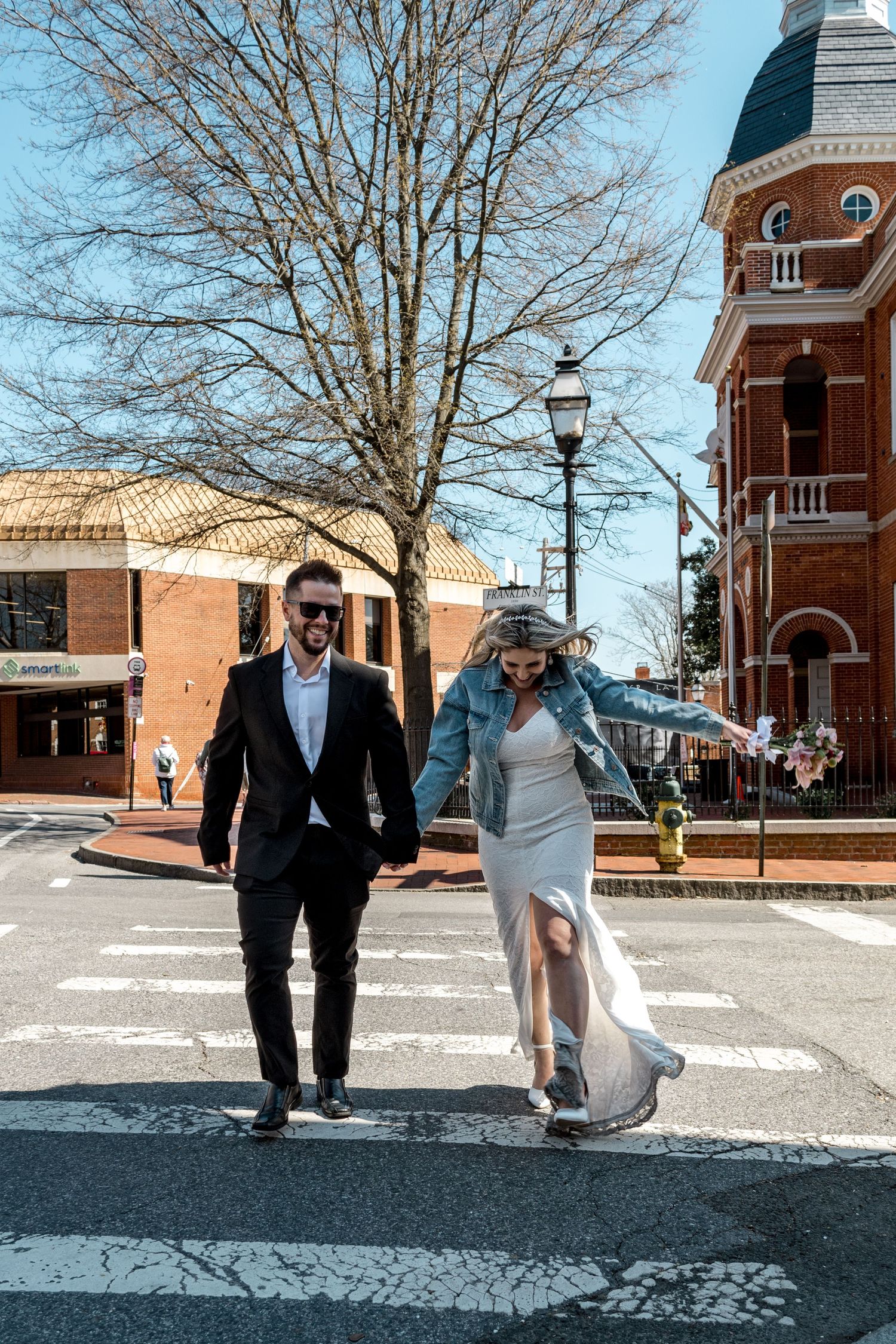Elegantly dressed couple crossing city street near historic brick architecture.