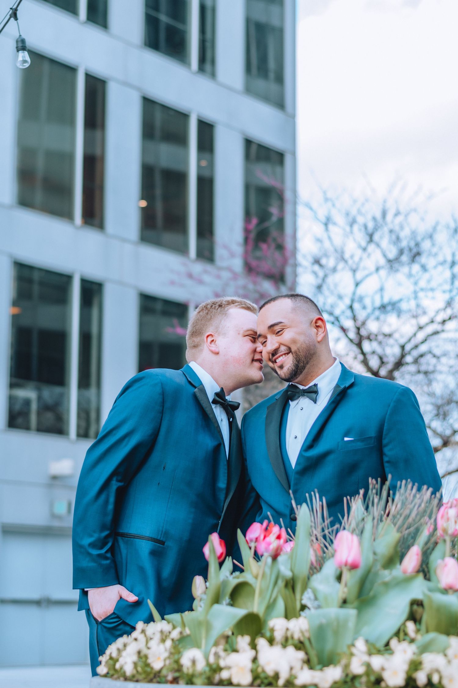 Two people share an intimate outdoor moment in front of a modern building with spring flowers blooming in the foreground.