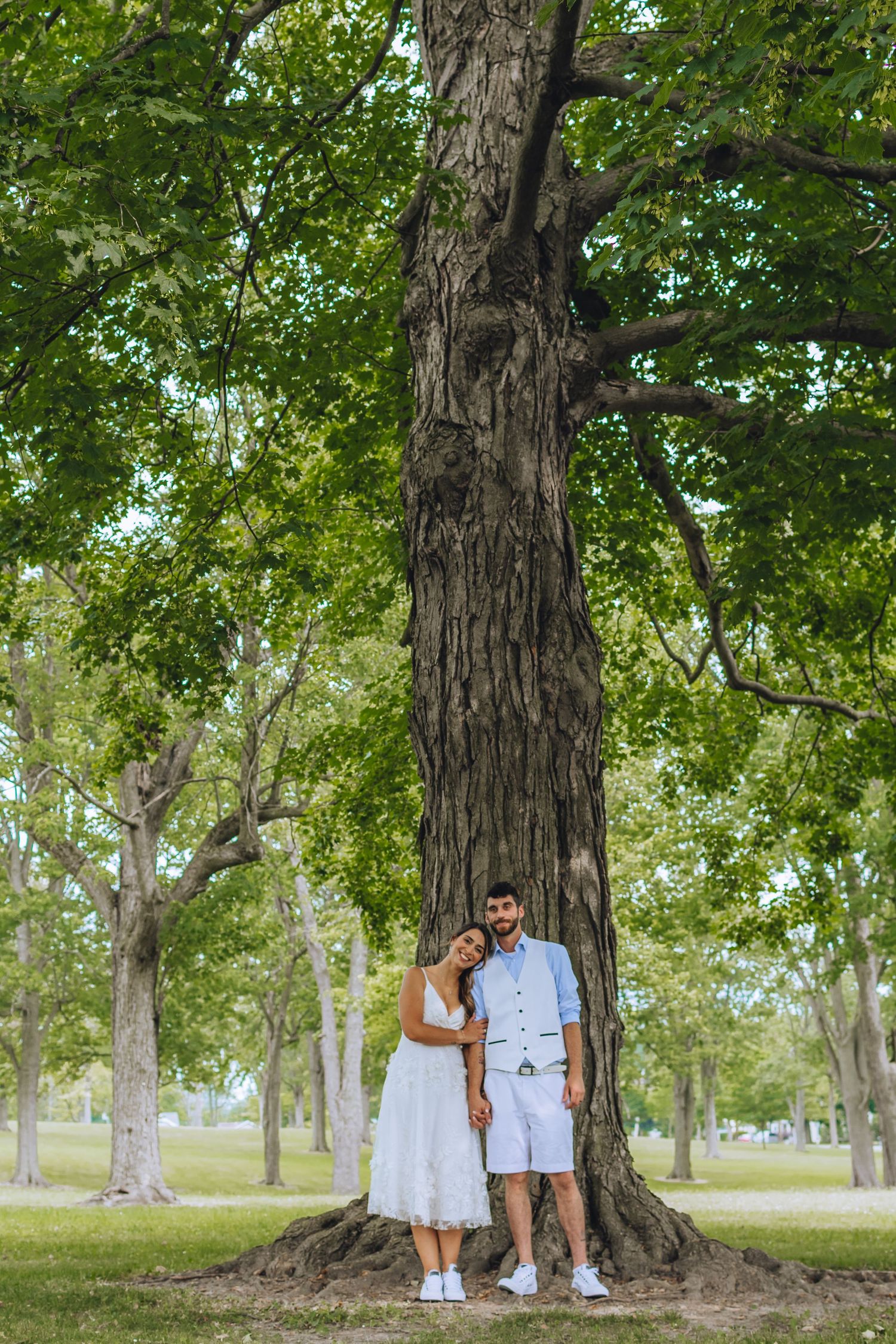 A couple in matching white outfits poses together near a large tree trunk in a peaceful park setting.