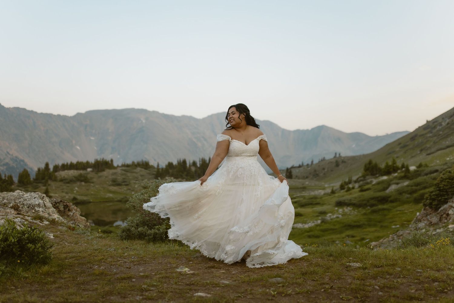 In the mountains at sunset, a bride in a flowing white gown sits among the rugged alpine landscape.