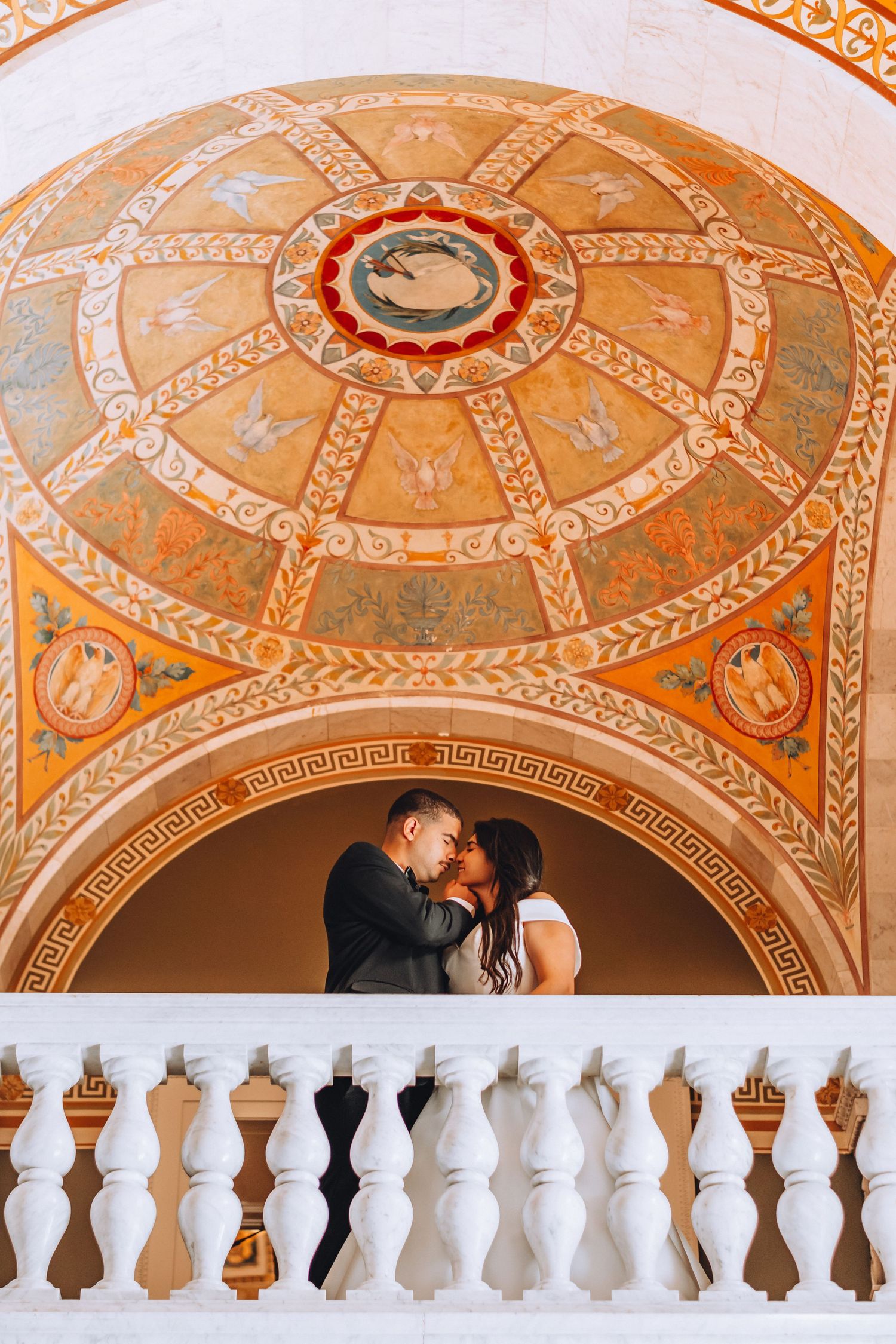 Newlyweds share a romantic moment under an ornately decorated dome ceiling with geometric patterns.