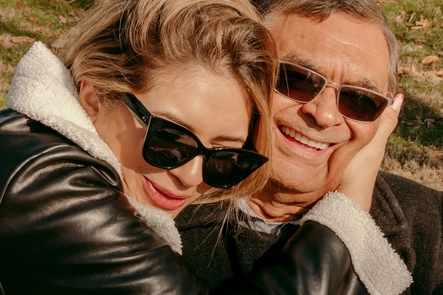 Two people wearing sunglasses share an intimate moment while wearing leather and sherpa jackets outdoors.