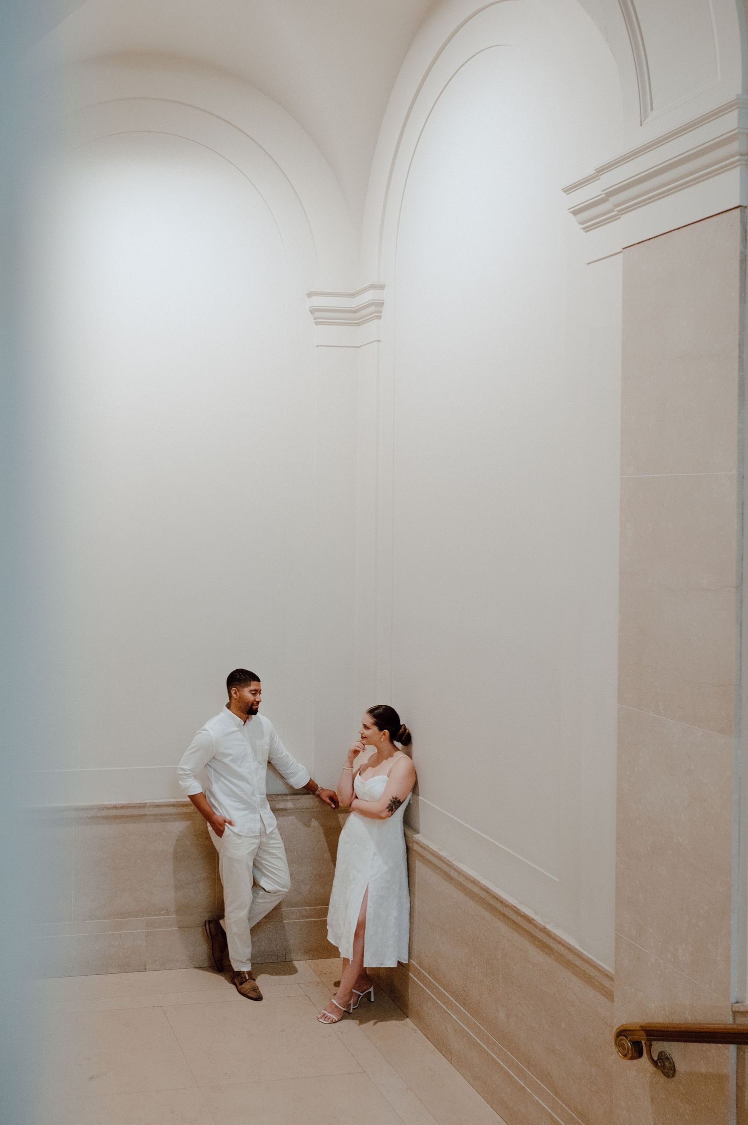 A couple shares an intimate moment in a bright, minimalist hallway with white walls and arched architectural details.