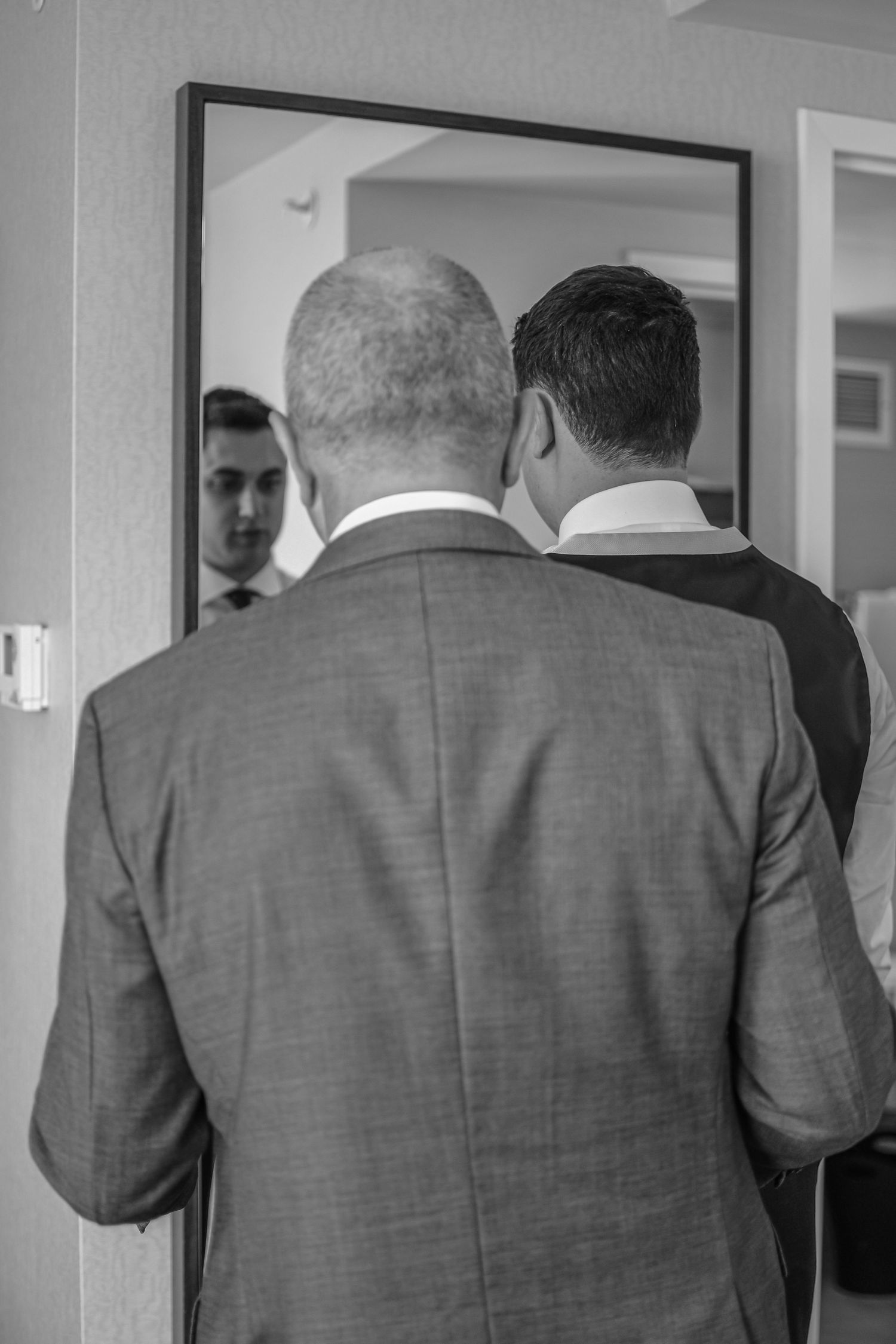 Black and white view of two men in suits looking into a mirror reflecting their formal attire.