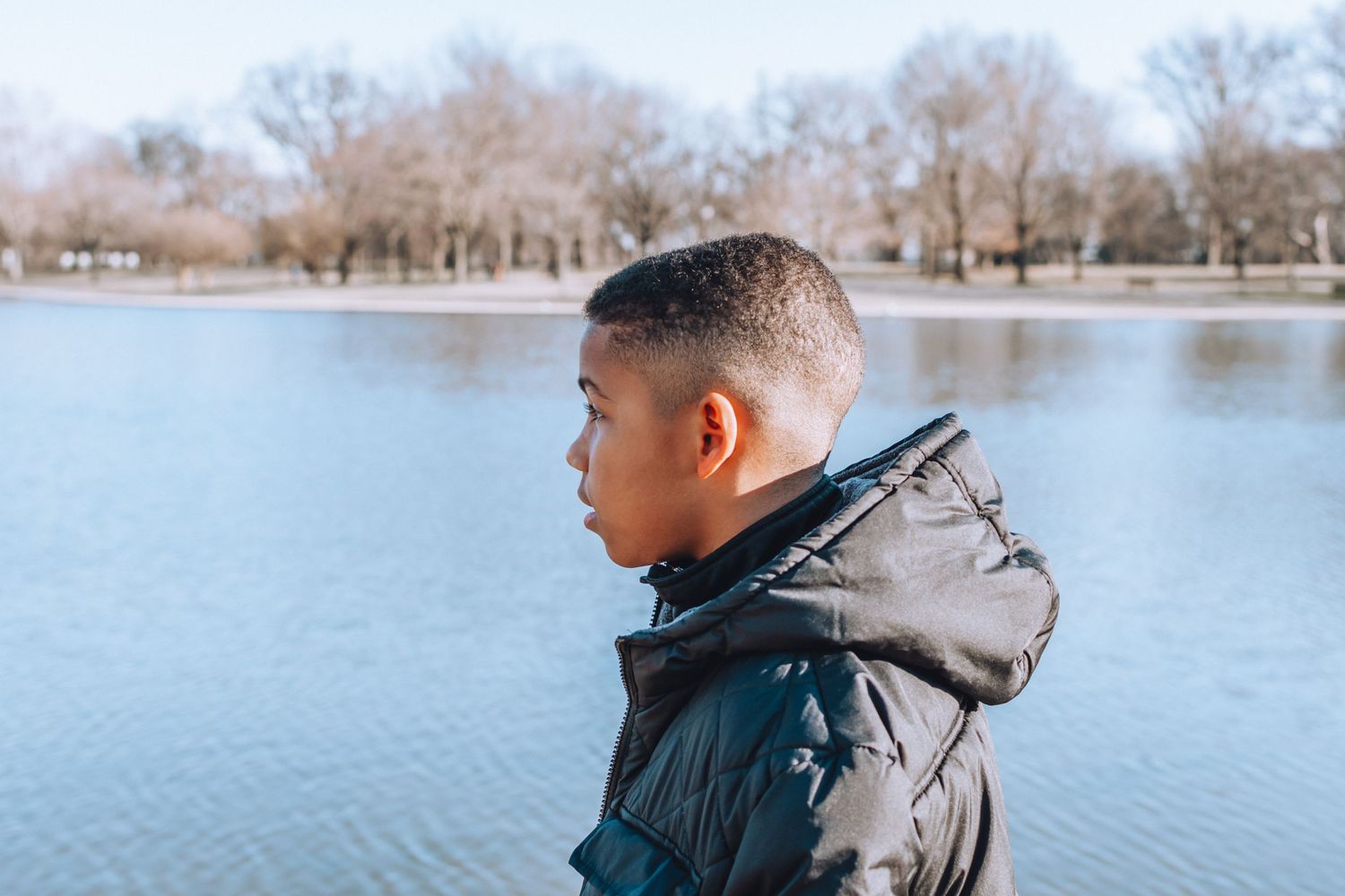Person in a gray winter jacket looks contemplatively at a calm lake with bare trees in background.