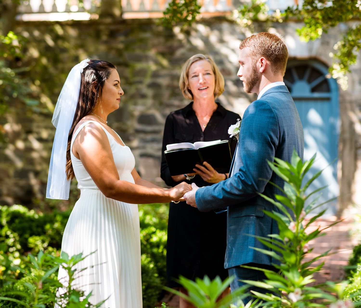 Intimate garden wedding ceremony with couple holding hands in front of stone building.