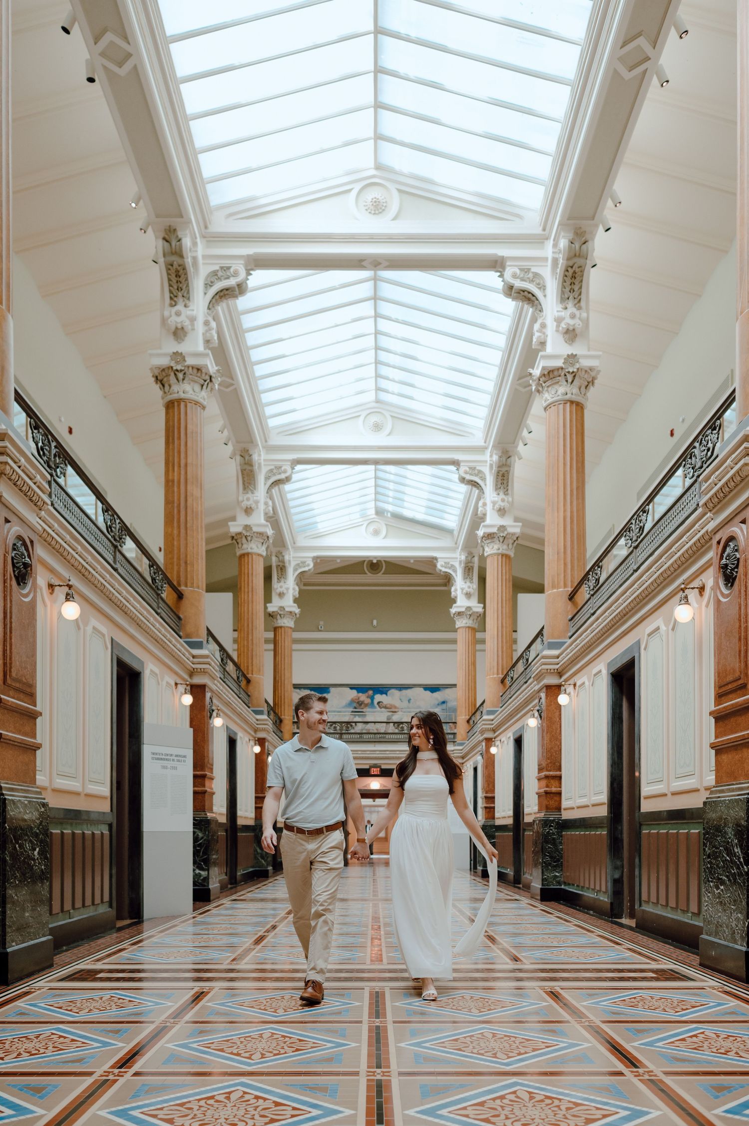 A couple walks through an elegant corridor with ornate columns, skylight ceiling, and decorative marble flooring.