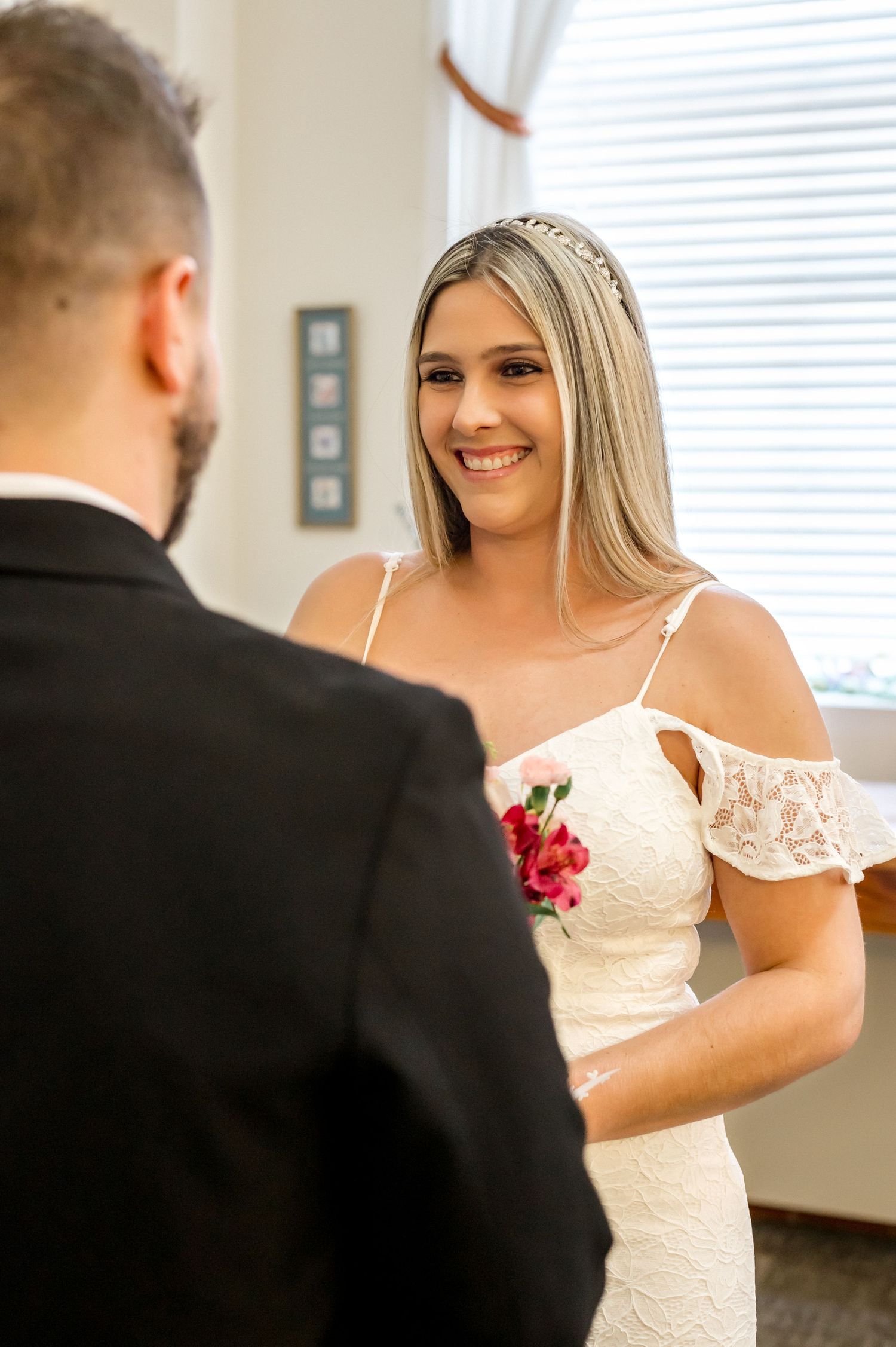 A bride in an elegant white dress with off-shoulder detail smiles warmly during the wedding ceremony.