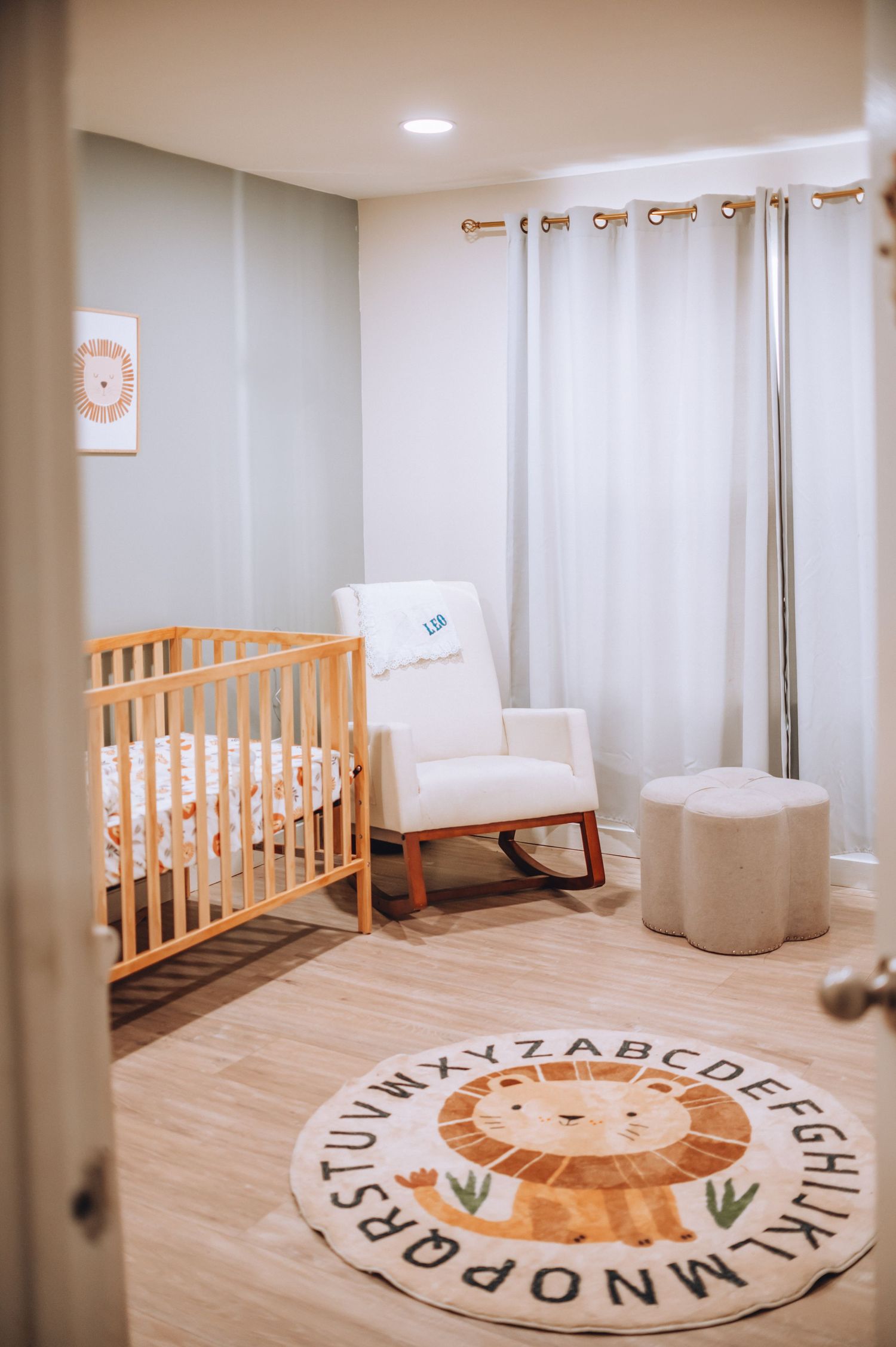 Neutral nursery with wooden crib, rocking chair, and circular lion alphabet rug.