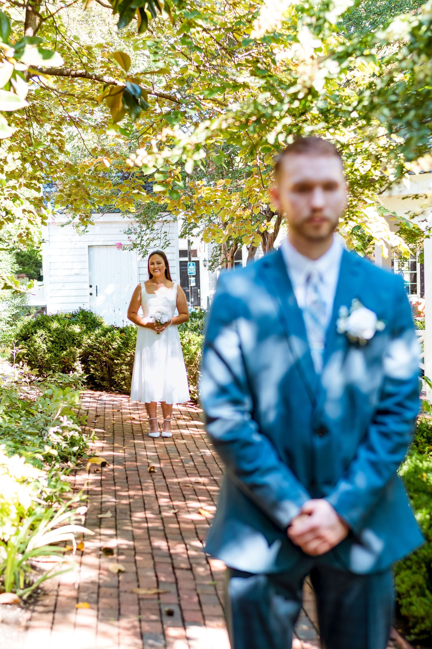 Wedding first look moment captured on brick pathway with greenery and white building in background.