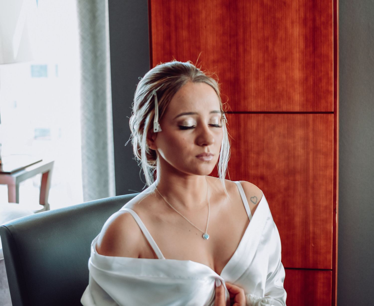 Woman with eyes closed wearing white off-shoulder top and silver necklace sits peacefully in soft lighting.