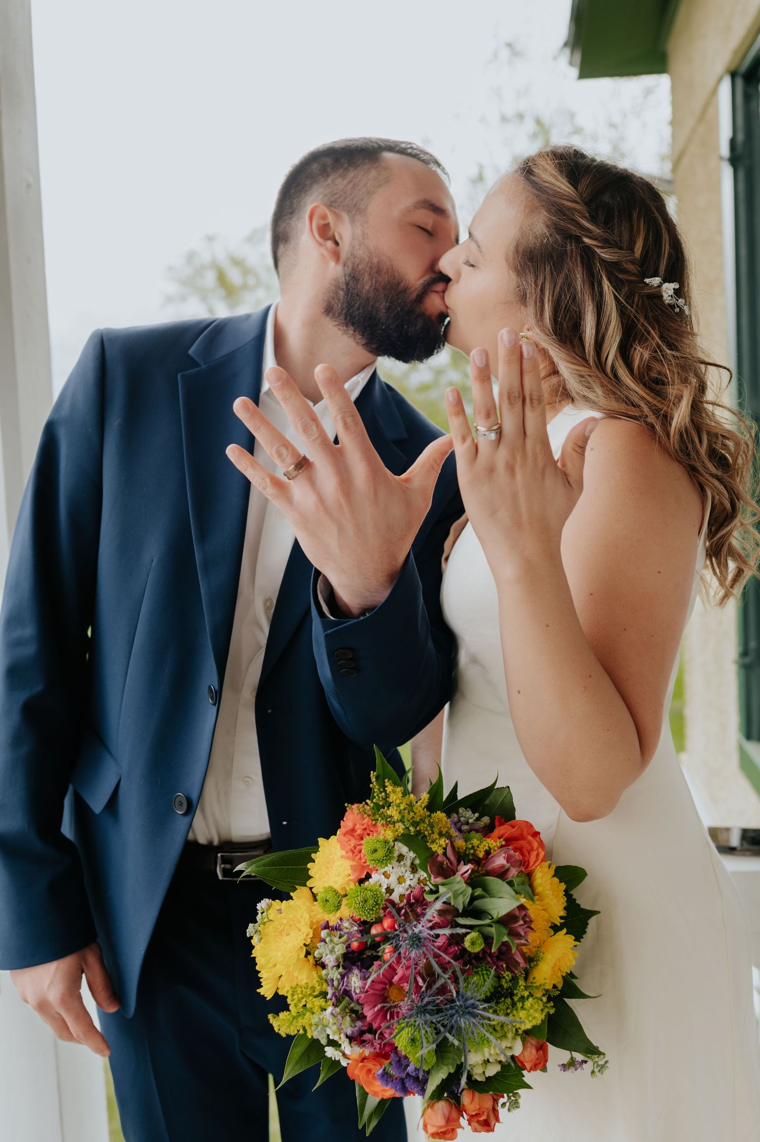 A newlywed couple kissing while showing off their rings and holding a colorful wildflower bouquet.