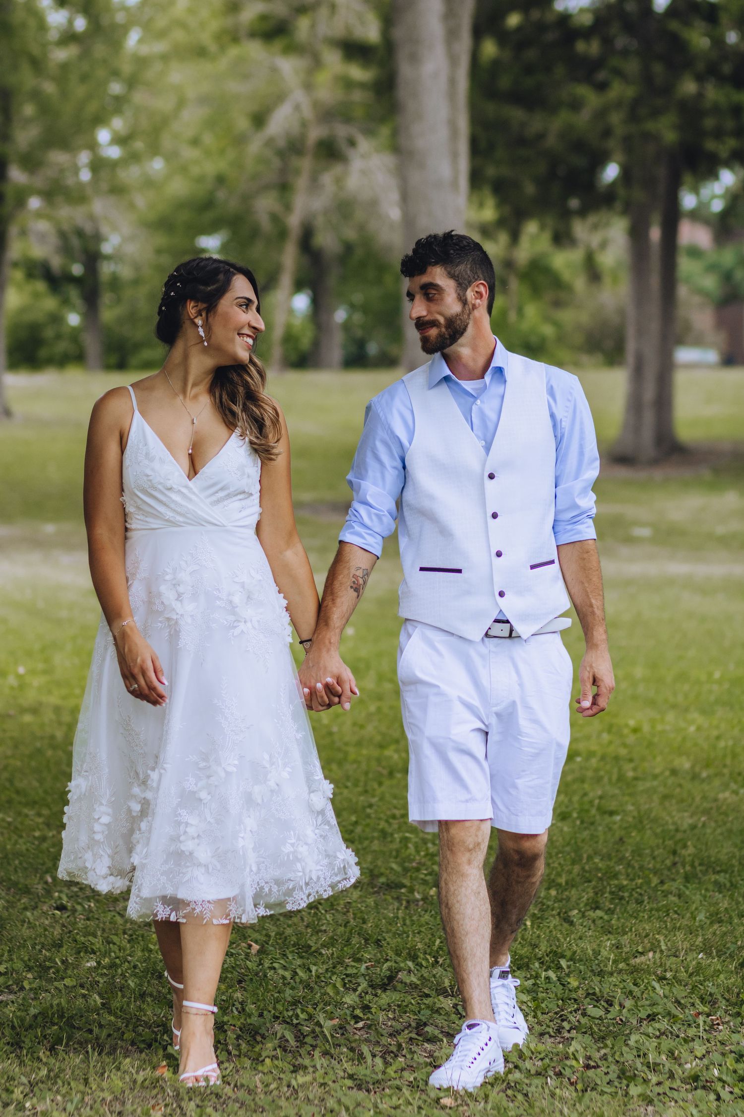 A couple walks hand in hand outdoors, with the woman wearing a white lace dress and the man in casual white attire.