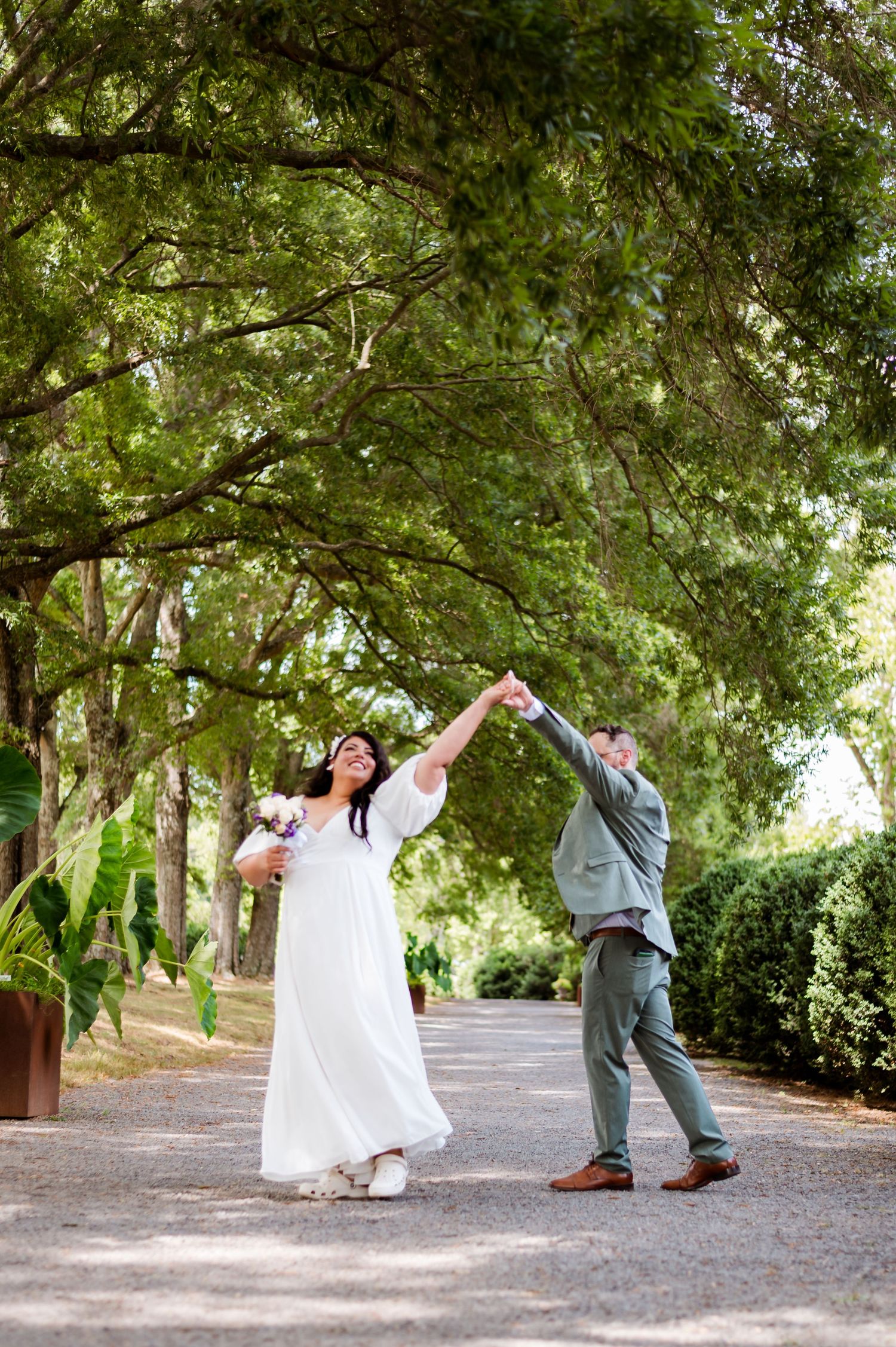 Newlyweds dancing joyfully on a tree-lined path in a garden setting.
