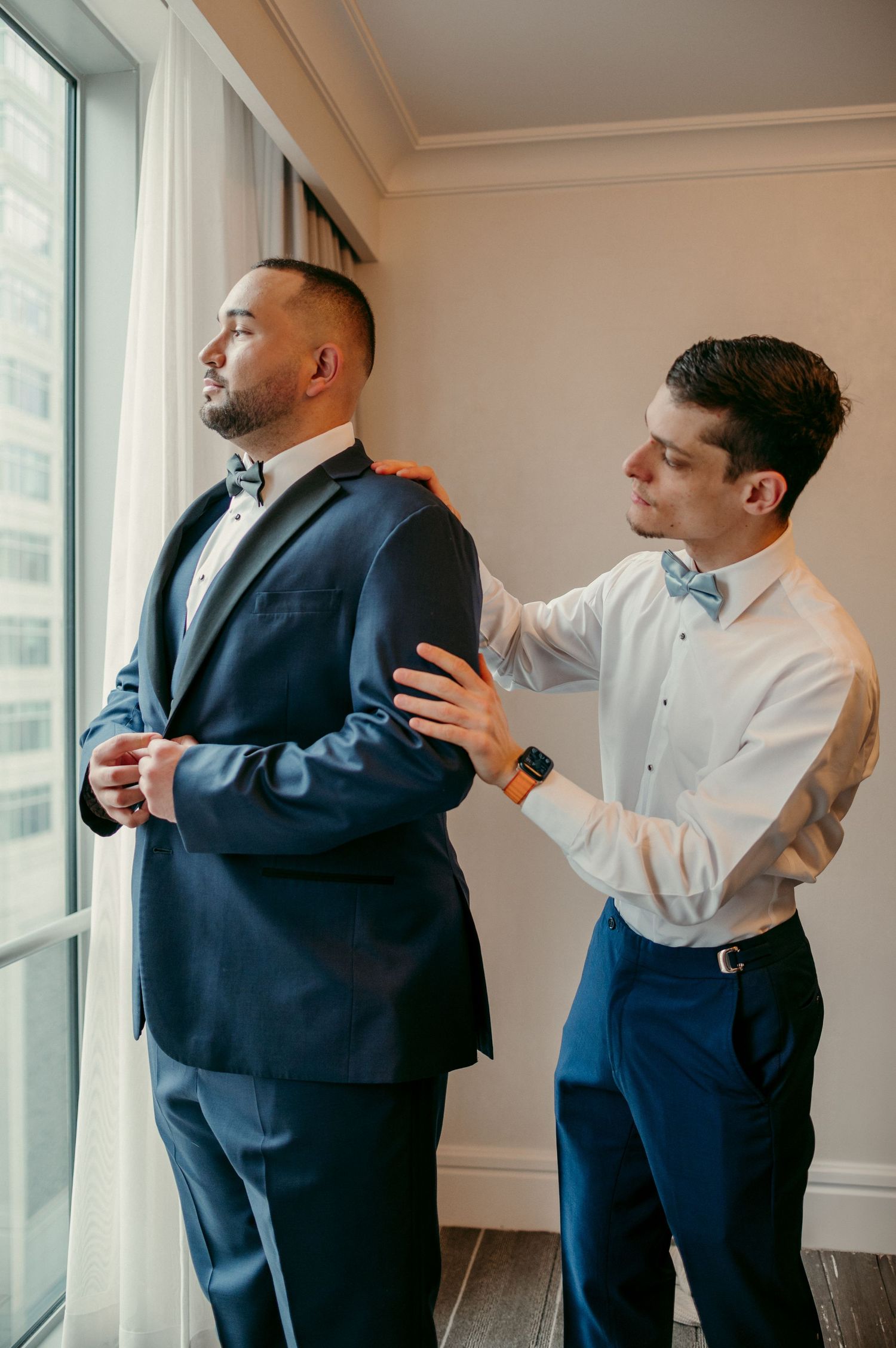 A sequence of photos showing two men helping adjust a navy suit jacket in a bright modern room with windows.