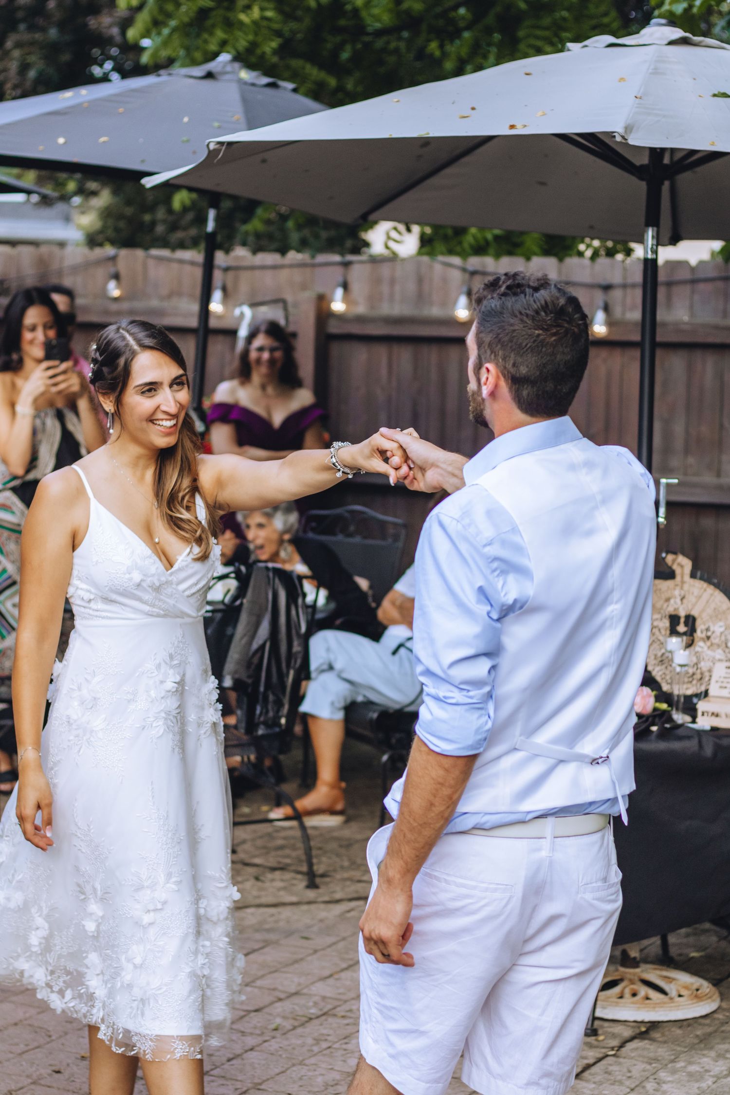 A couple dances together at an outdoor celebration, with both dressed in white attire on a paved patio.