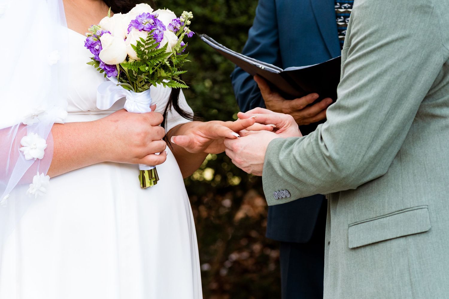 Hands exchanging rings during outdoor wedding ceremony with purple and white bouquet.