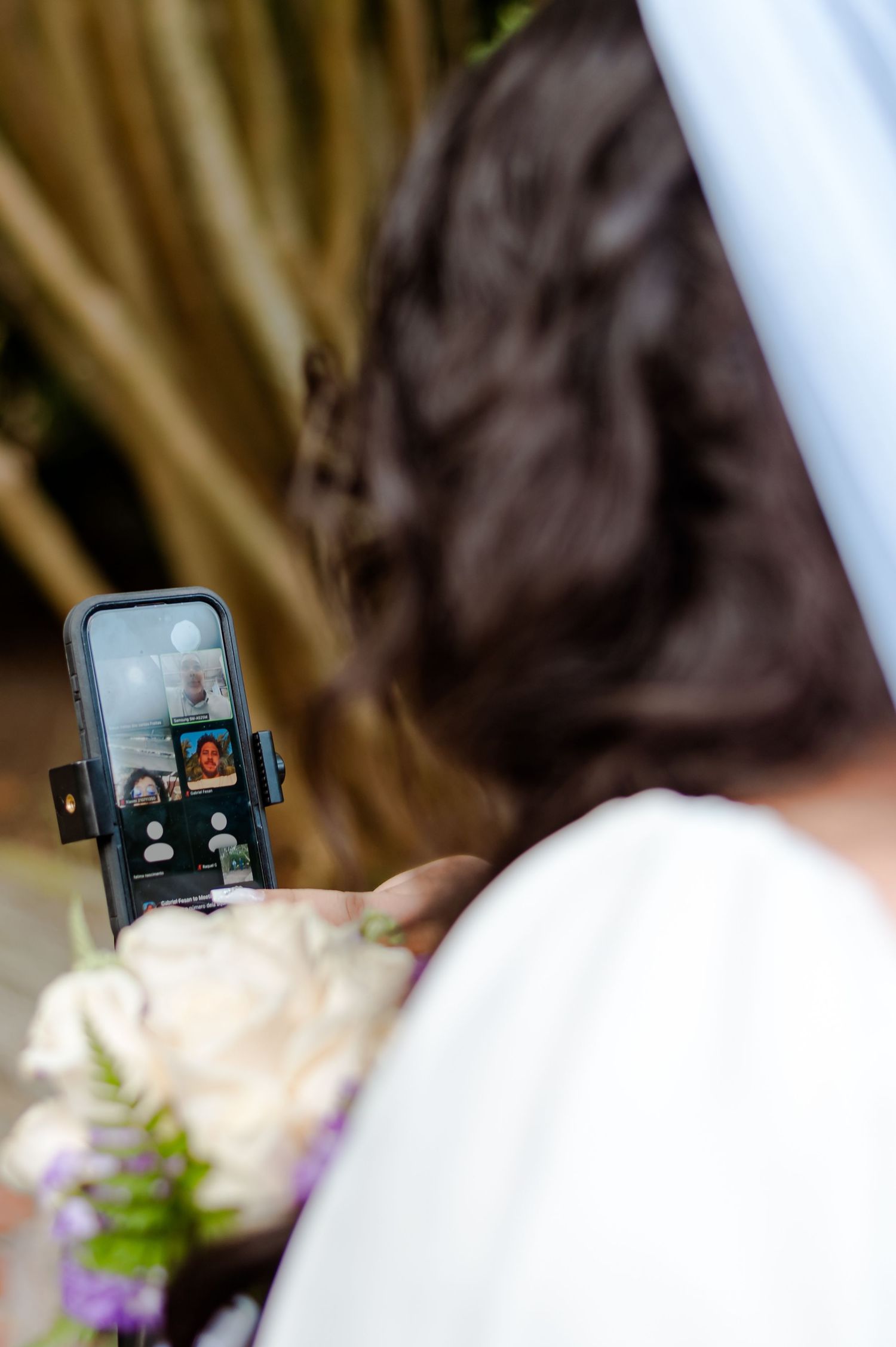 Smartphone displays video call on screen during wedding ceremony with bouquet in foreground.