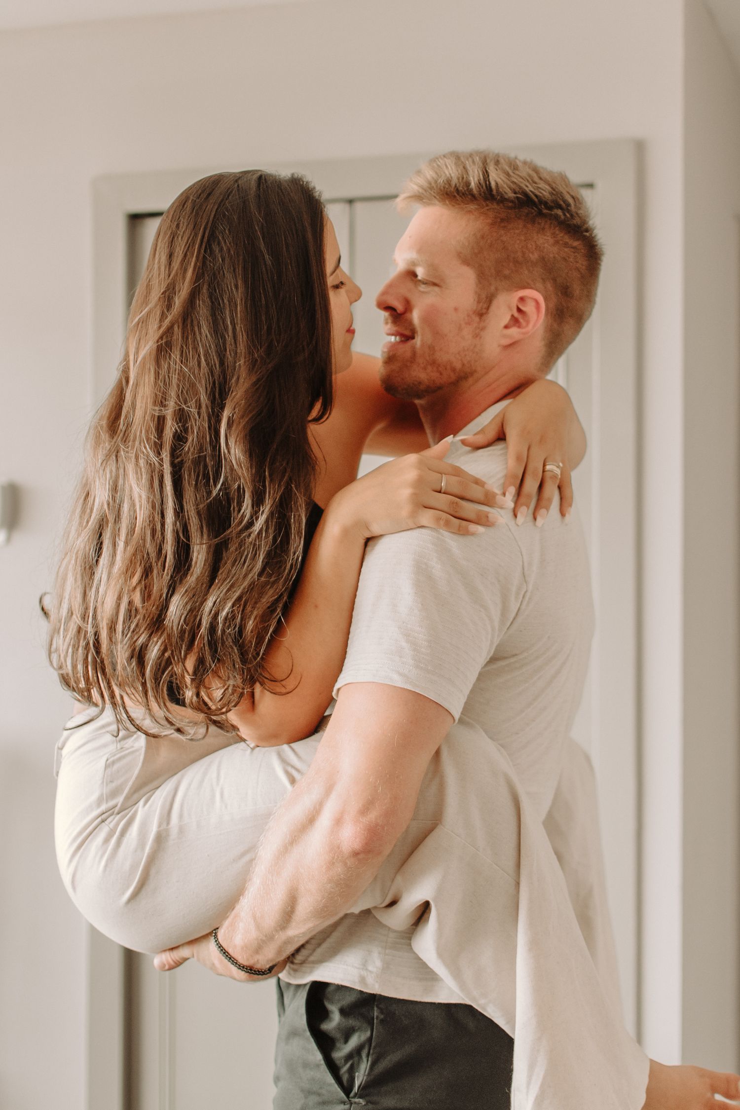 A romantic couple shares an intimate embrace against a white wall in casual clothing.