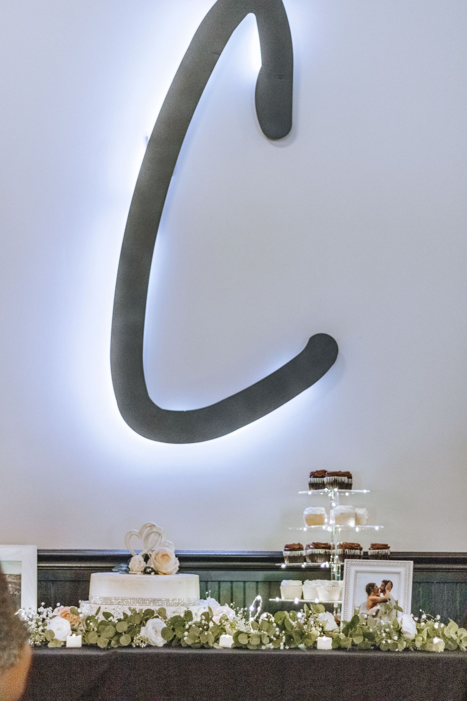 Wedding reception setup with large illuminated letter C on wall above floral decorations and dessert display table.