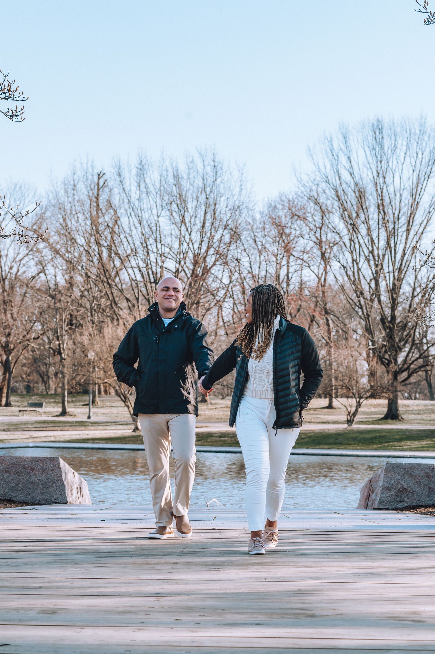Couple walking together on path near water with bare winter trees in background.