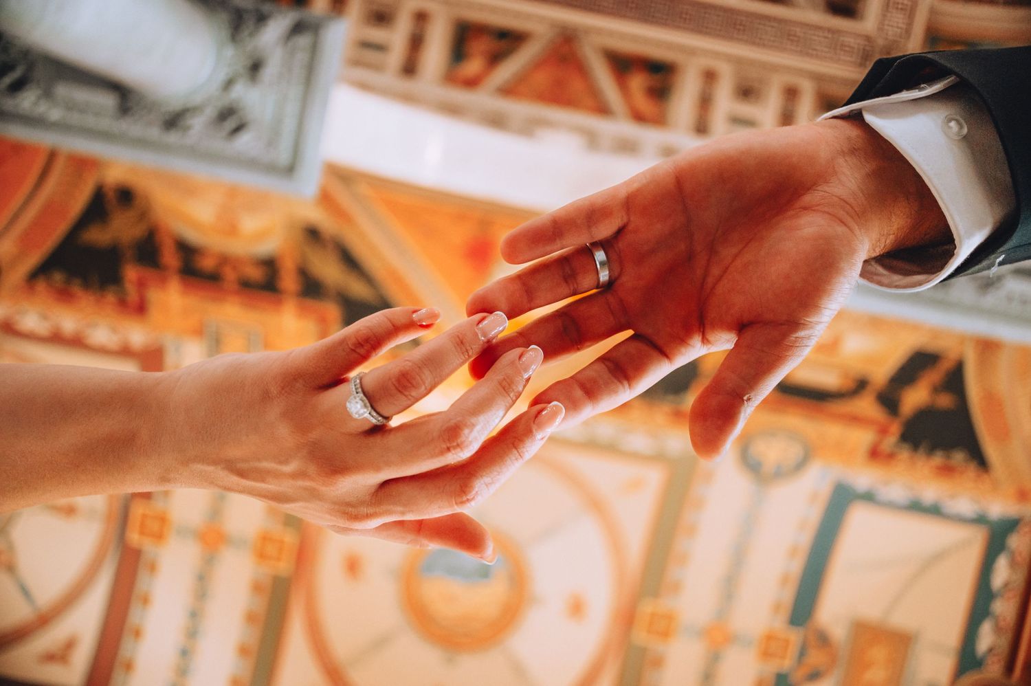 Close up of hands exchanging wedding rings during marriage ceremony with ornate ceiling visible in background.
