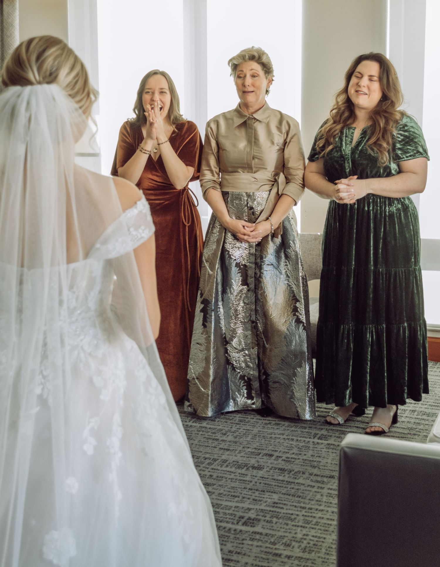 Three women in formal dresses react with joy upon seeing a bride in her wedding gown.