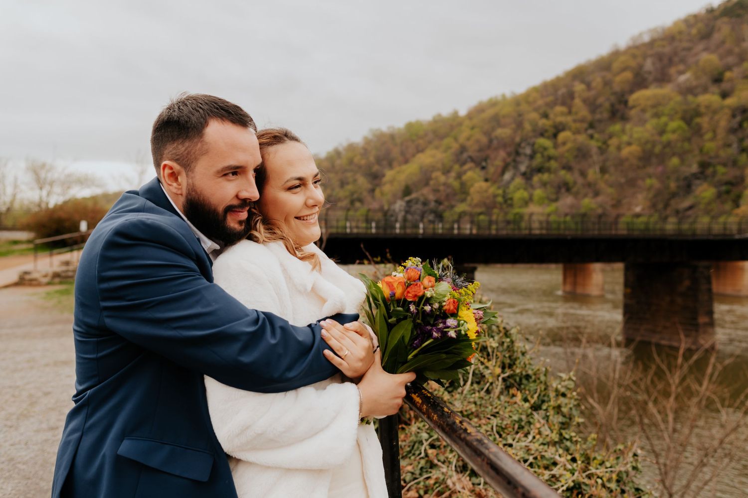 A joyful couple embraces outdoors near a rustic bridge on an autumn day, with the bride holding a vibrant floral bouquet.