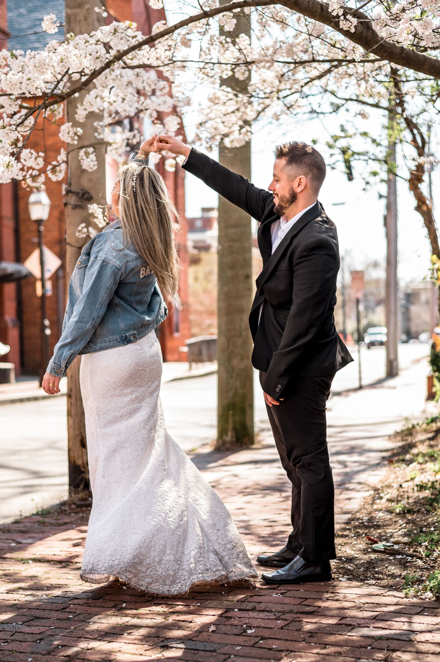 Couple posing under blooming cherry trees on brick sidewalk in wedding attire.