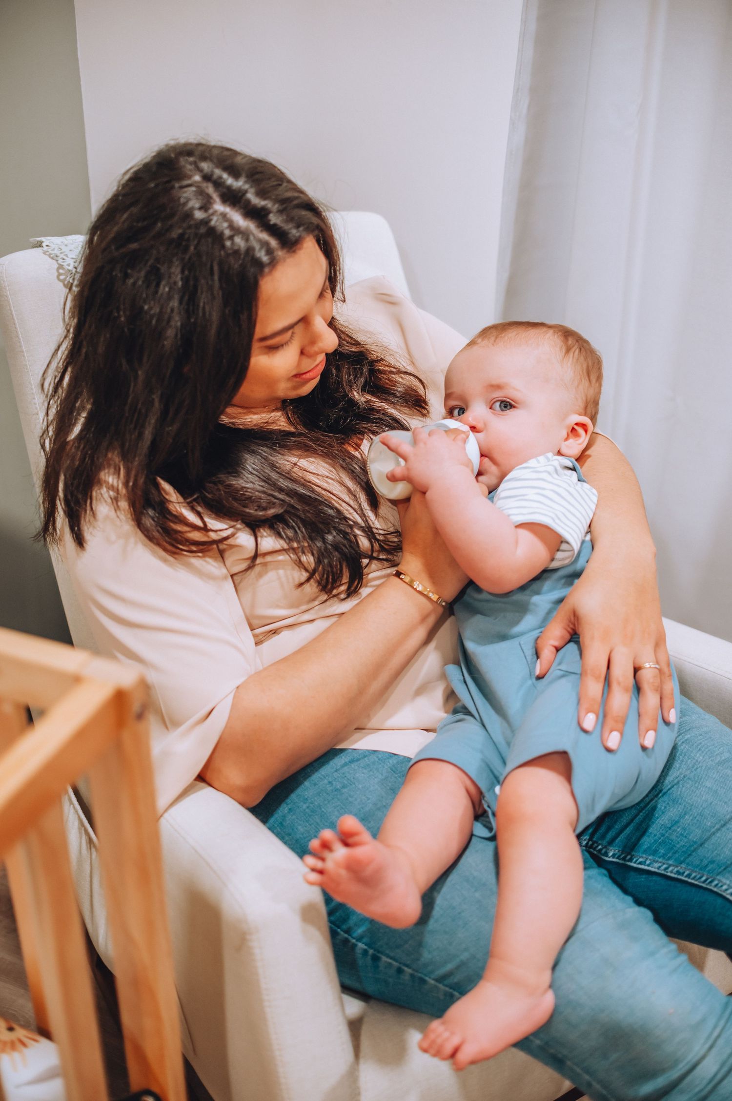 Tender moment of feeding time in white nursery chair with baby in blue overalls.