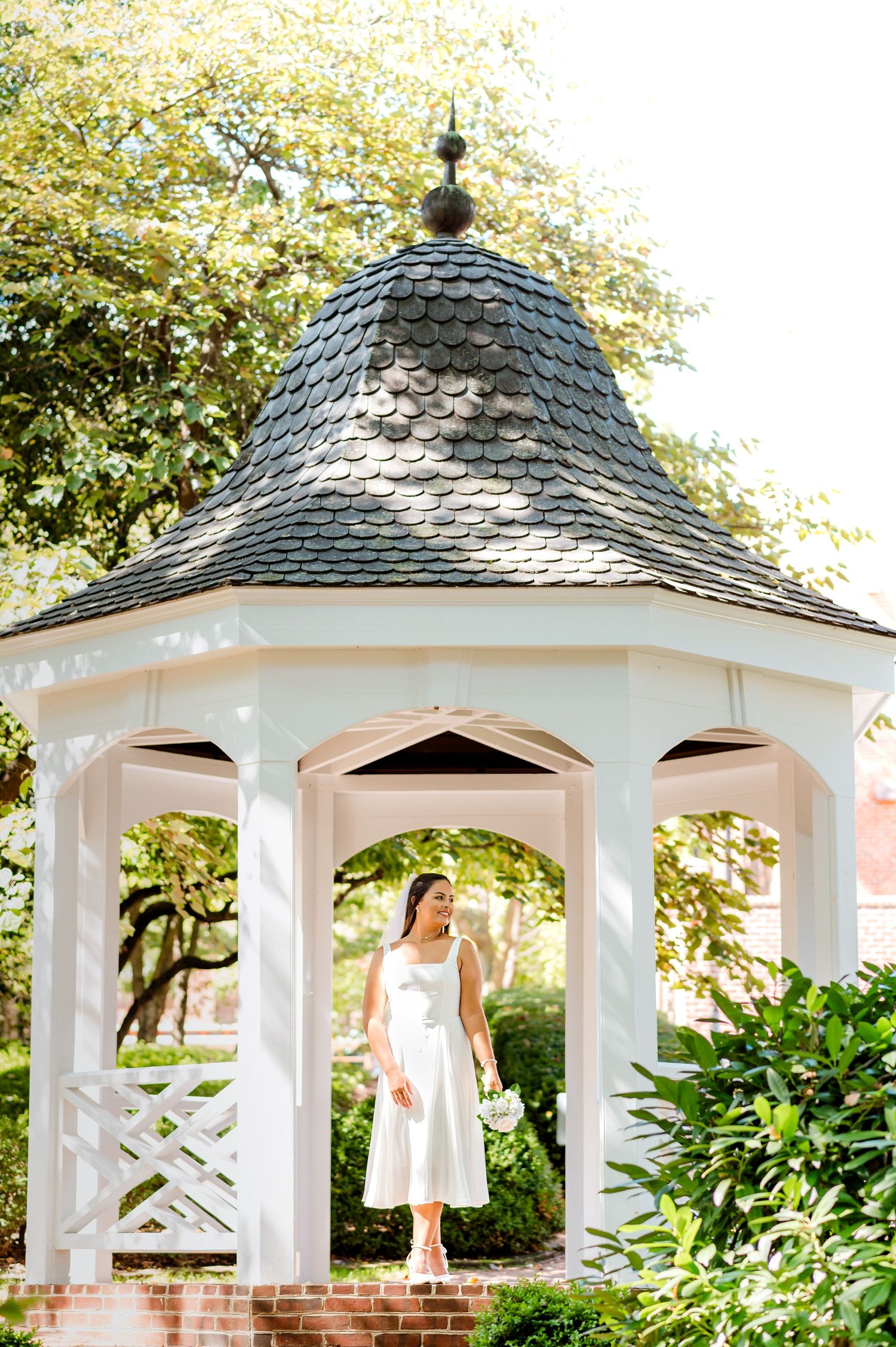 Bride in tea-length dress poses in classic white gazebo with shingled roof surrounded by greenery.