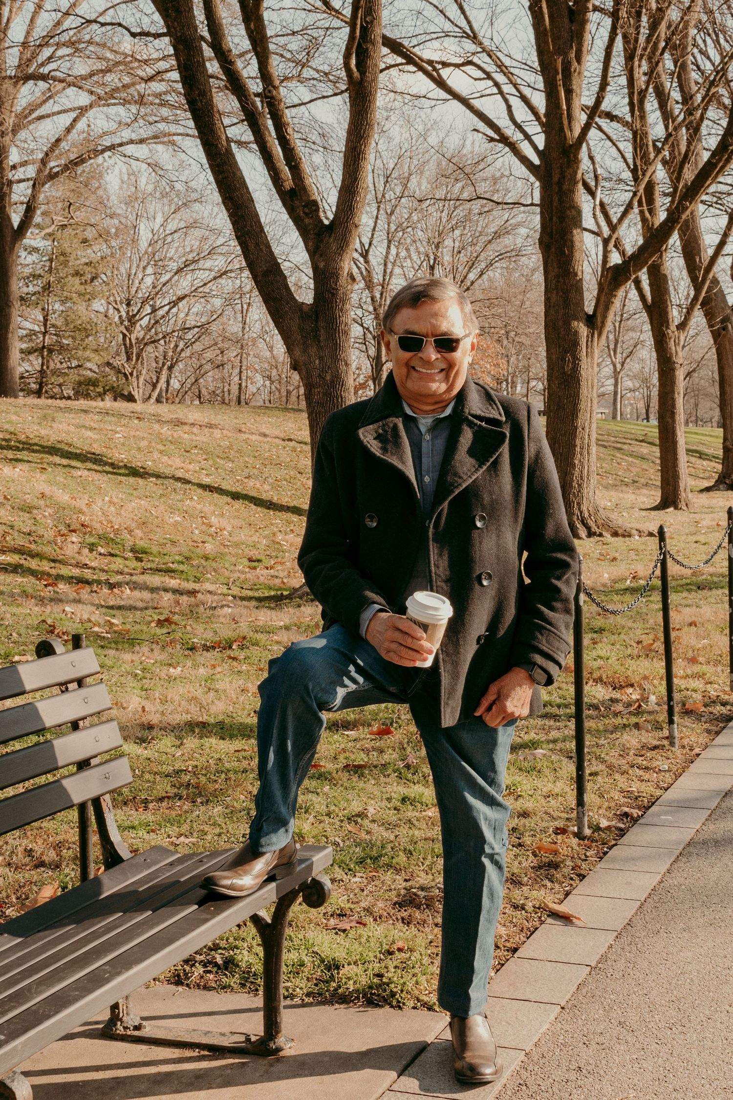 Person in sunglasses and dark coat sits casually on park bench holding coffee cup.