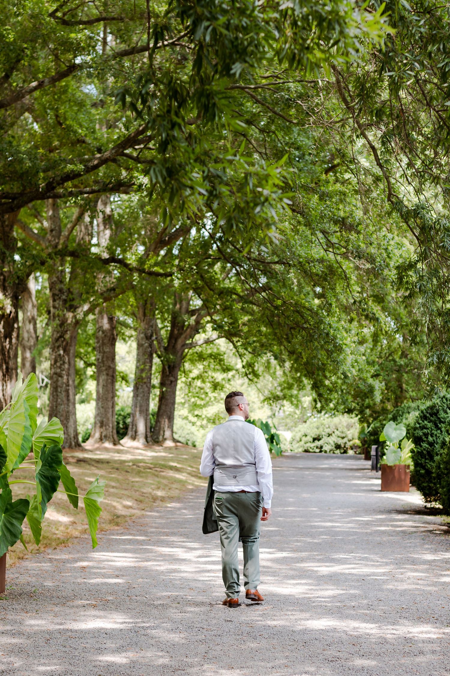 Two people walk along a shaded tree-lined path with dappled sunlight filtering through the green canopy above.