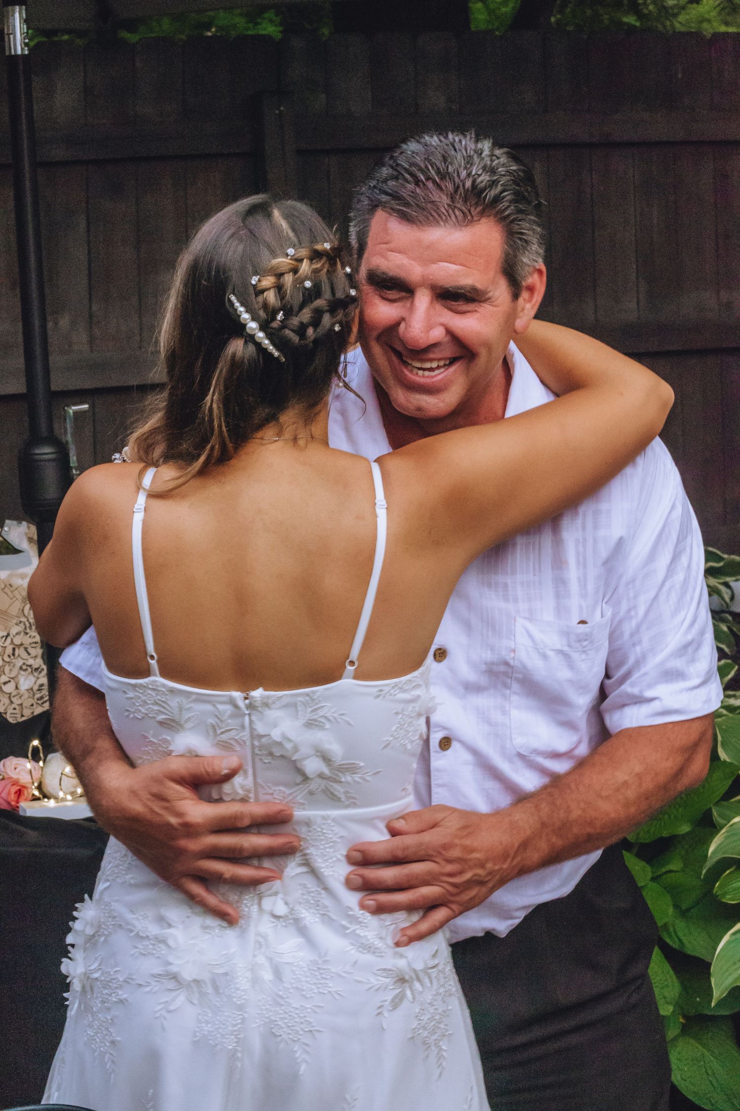 A joyful embrace between a bride in a white lace gown and wedding guest outdoors during a celebration.