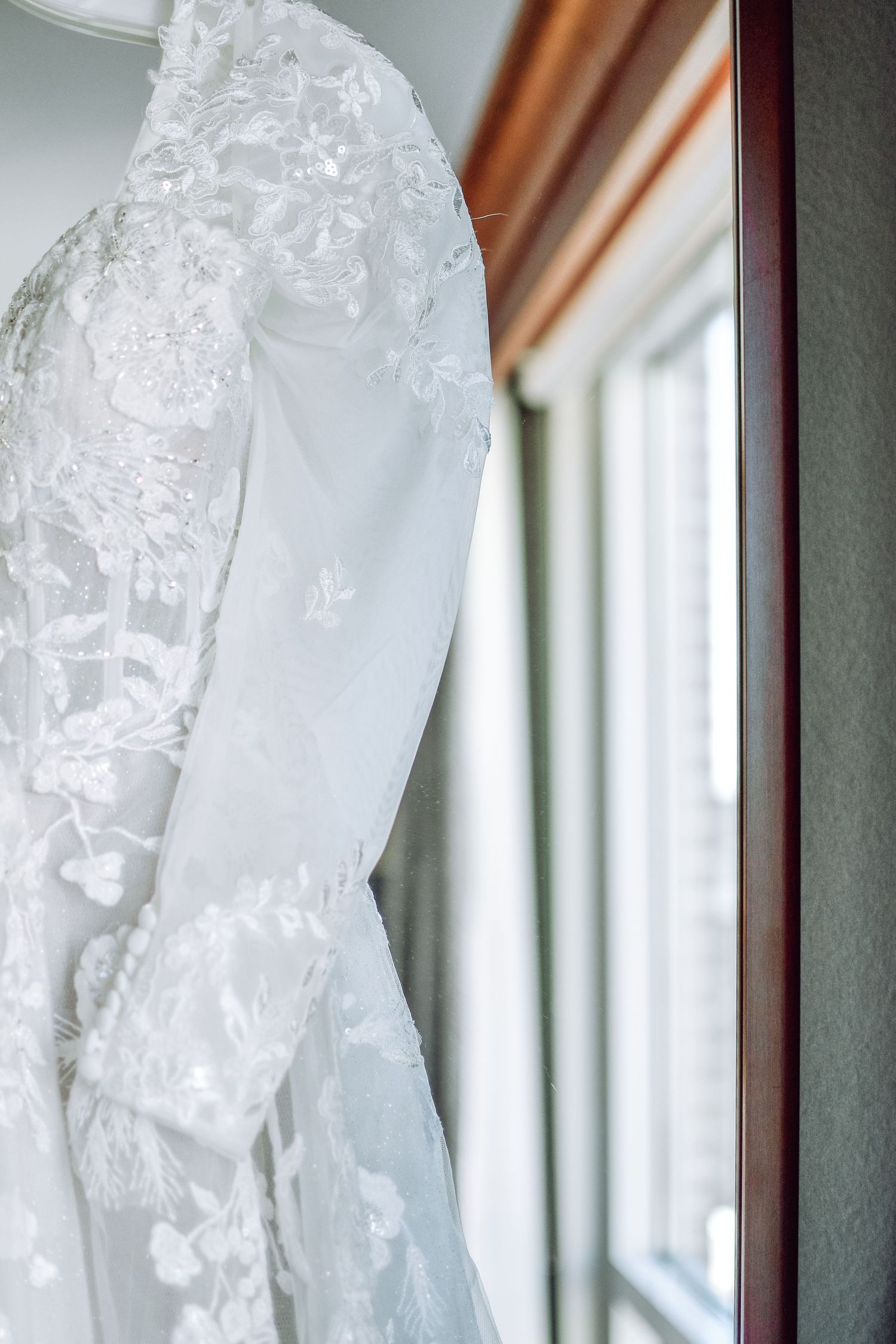 Wedding dress with delicate lace details hangs near a window in soft natural light.