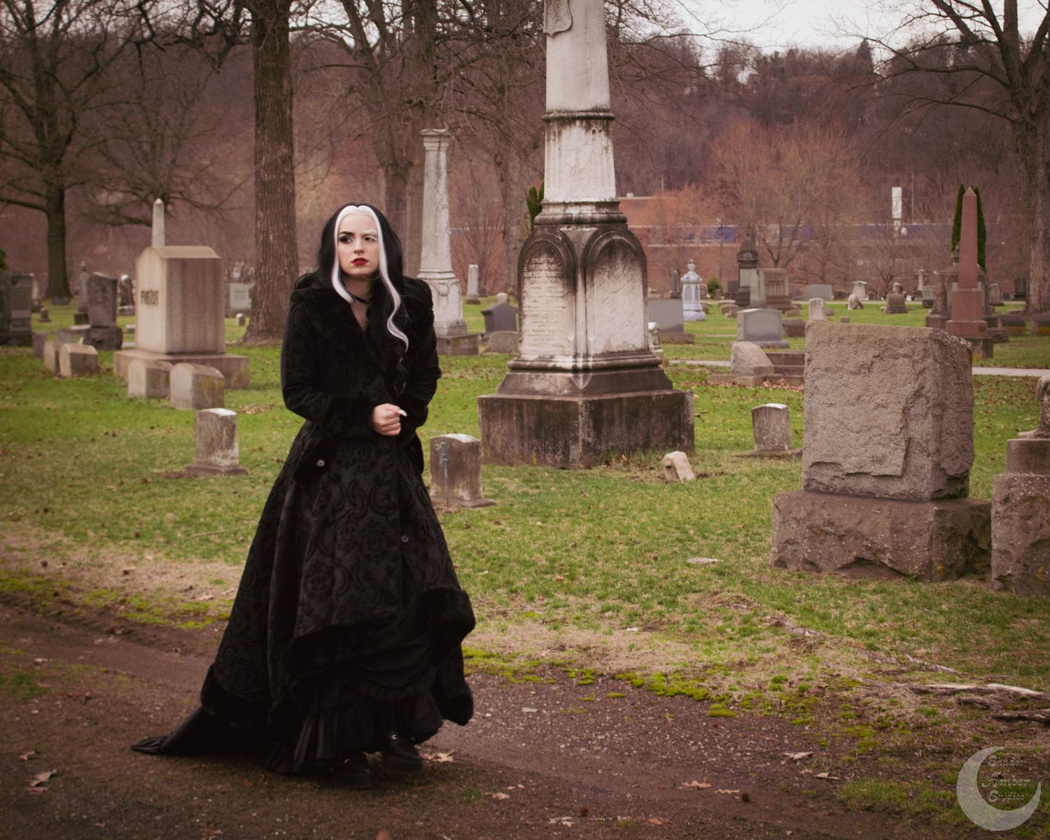Gothic woman in black dress and nun headpiece walking through an old cemetery with stone monuments.