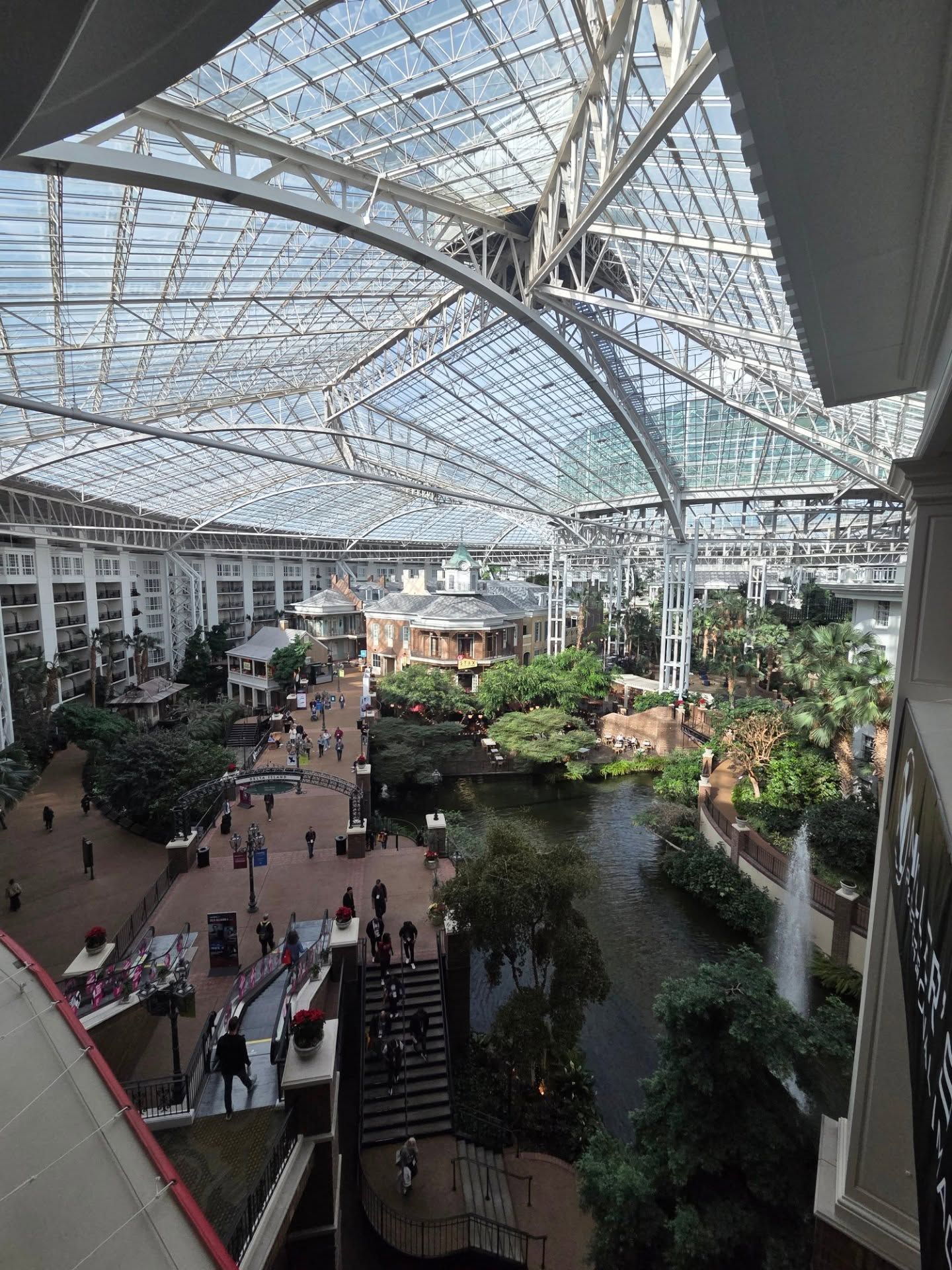 Large indoor atrium with glass ceiling, multiple levels, and tropical plants in a modern shopping mall or commercial space.