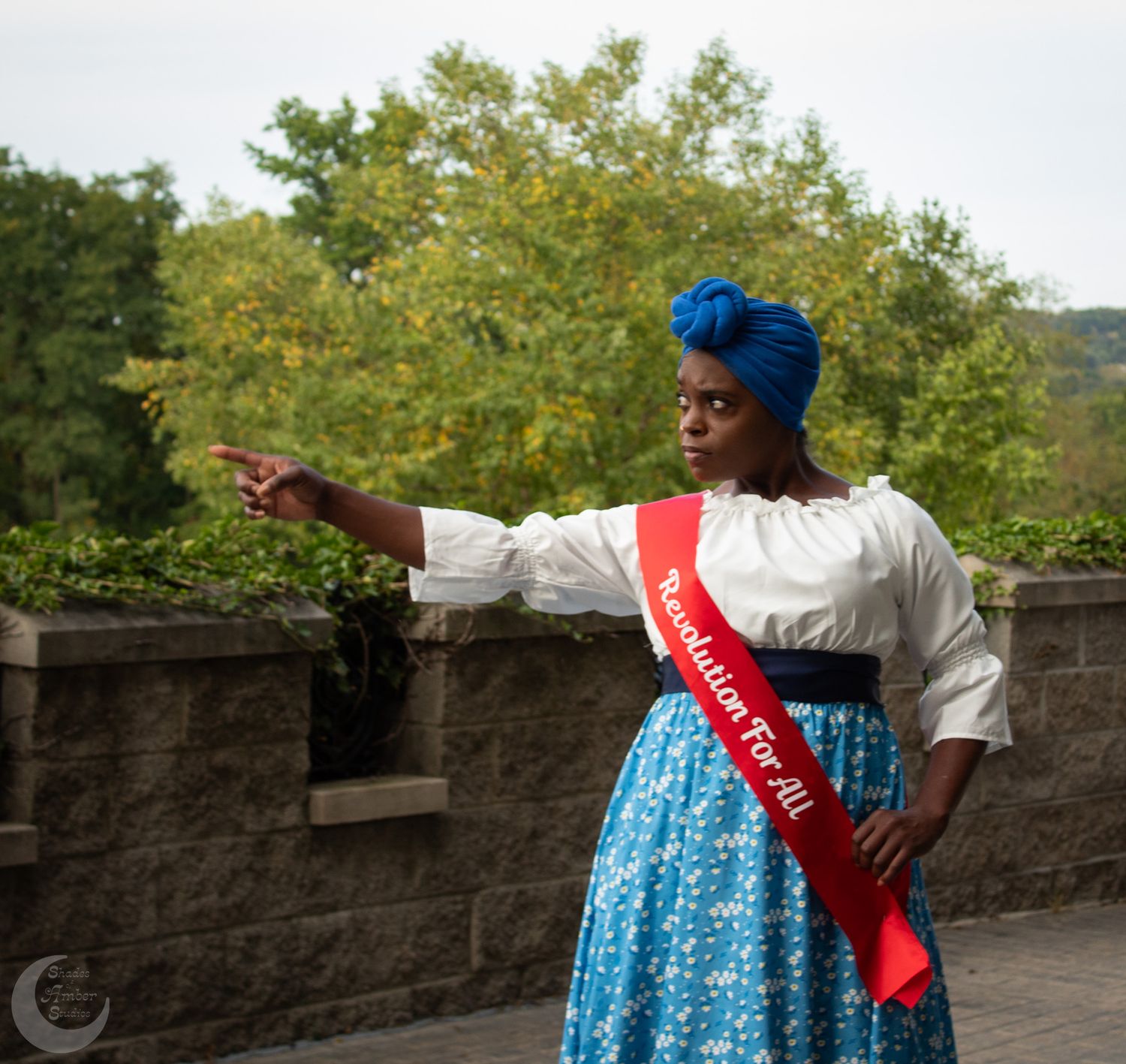 Costumed performer in colonial dress with blue headwrap and red sash gestures while standing near stone wall.