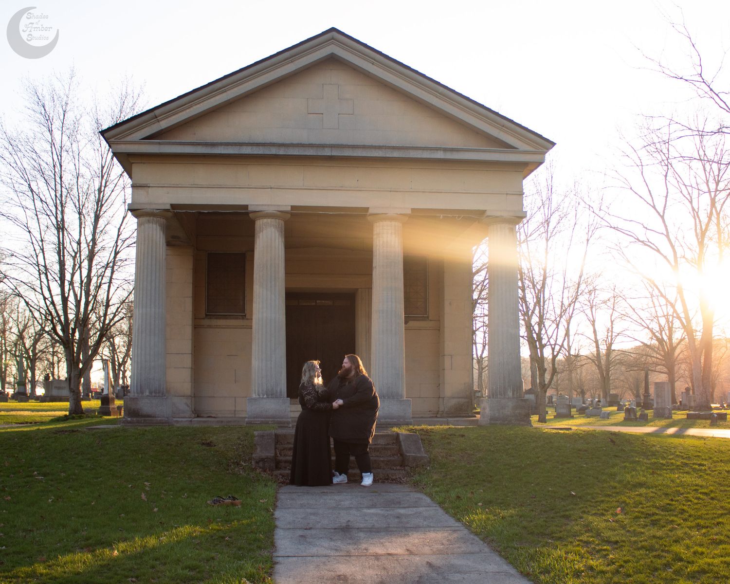 Couple in black attire standing before a neoclassical mausoleum at sunset in a cemetery.