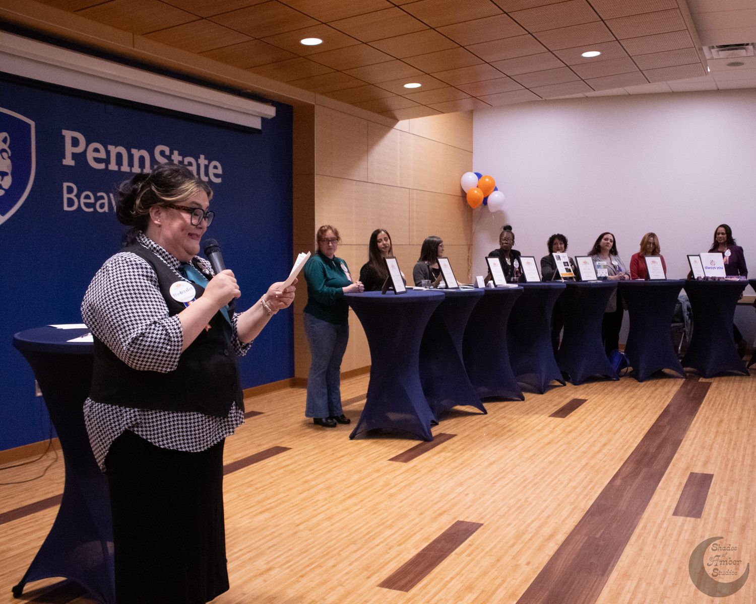 A presenter speaks into a microphone at a Penn State panel event while panelists sit at a long table on stage.