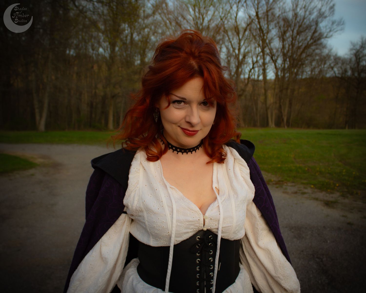 A woman with vibrant red hair wearing a corset and white blouse stands outdoors on a wooded walking path.