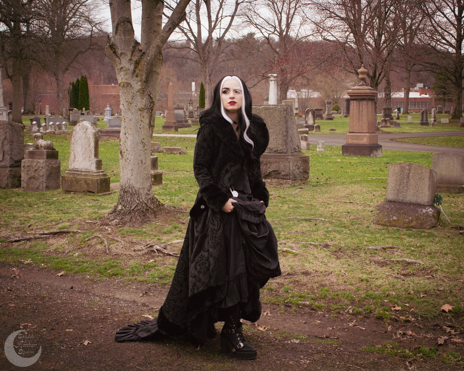 Gothic woman in black Victorian dress poses among gravestones in cemetery, featuring white hair and dramatic dark makeup.