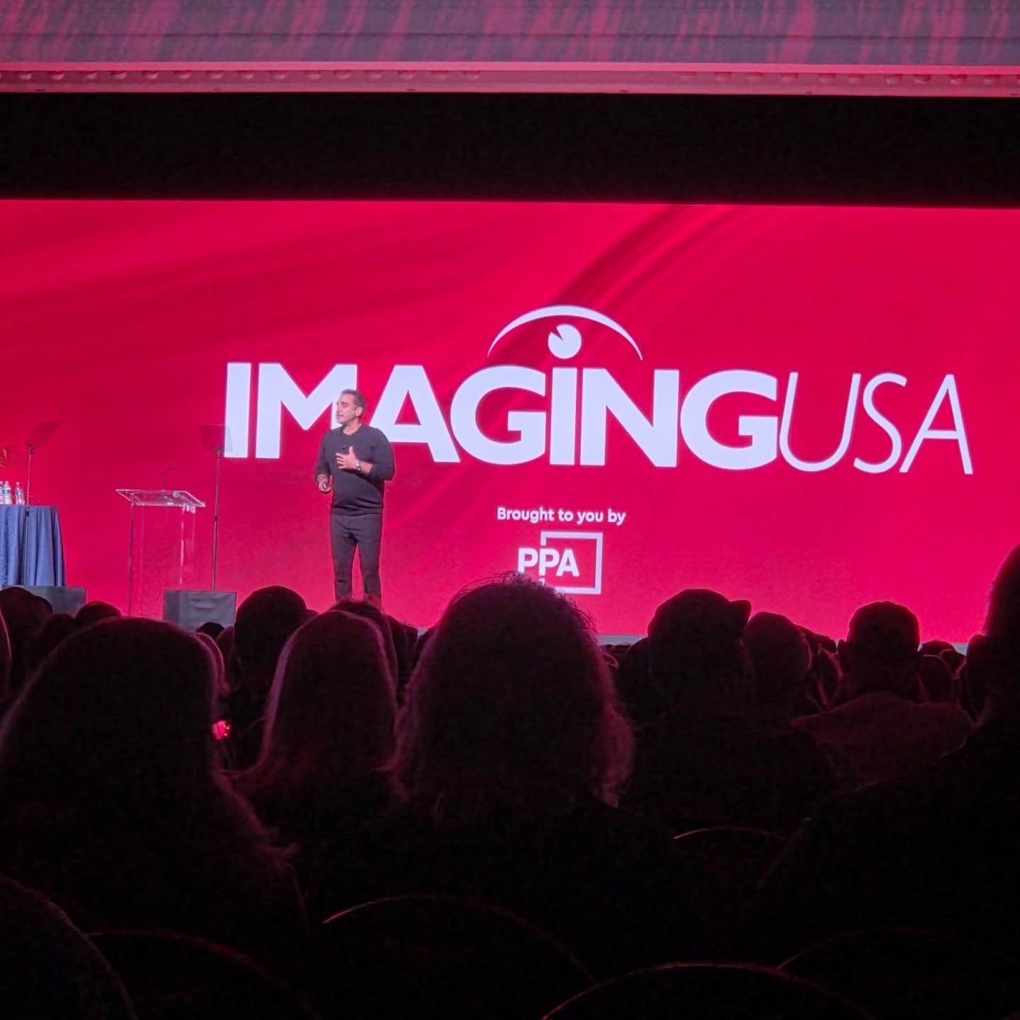 A presenter stands on stage at ImagingUSA conference with red backdrop and silhouetted audience in foreground.