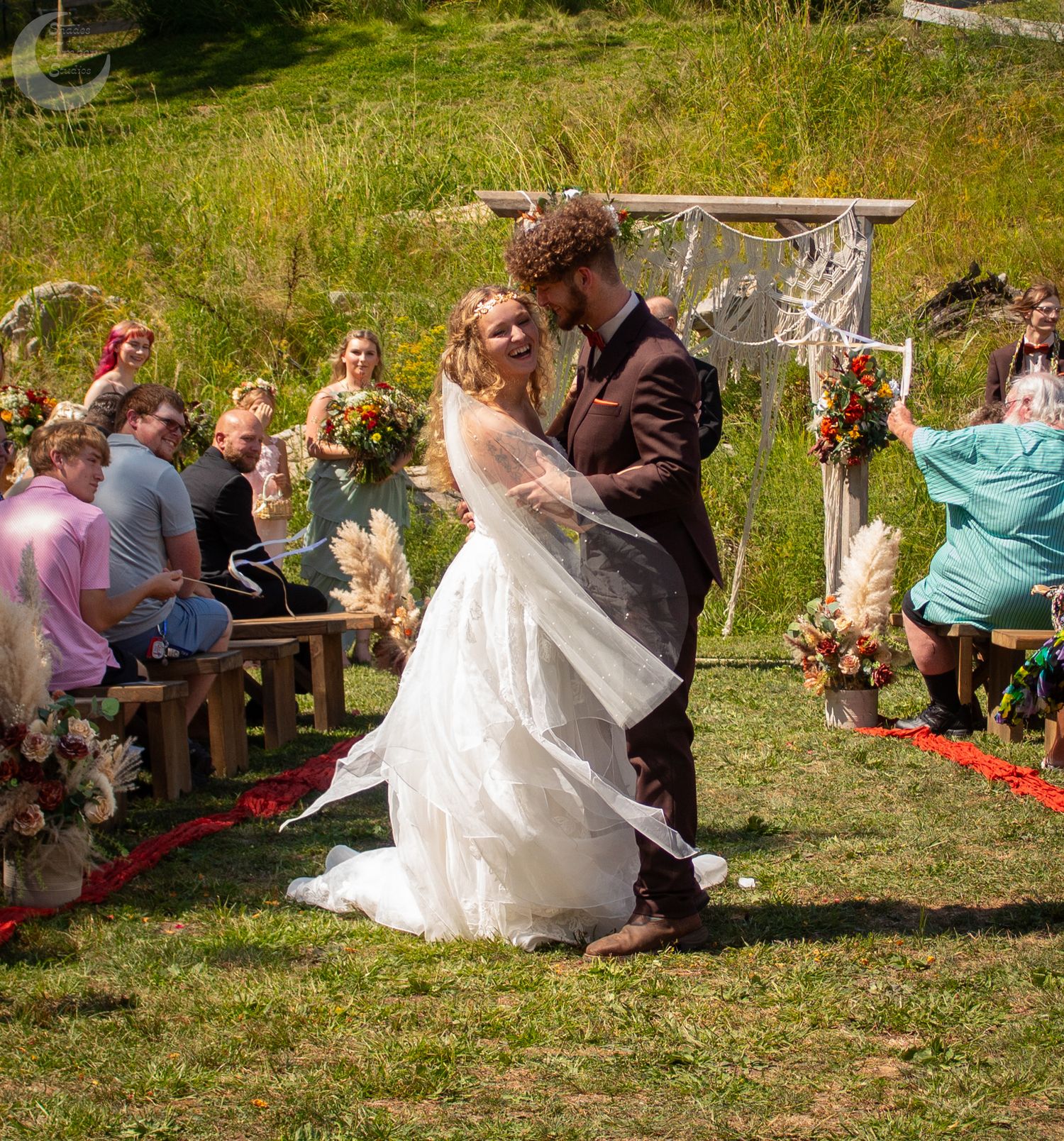 Outdoor wedding gathering with guests seated on benches in a grassy meadow setting.